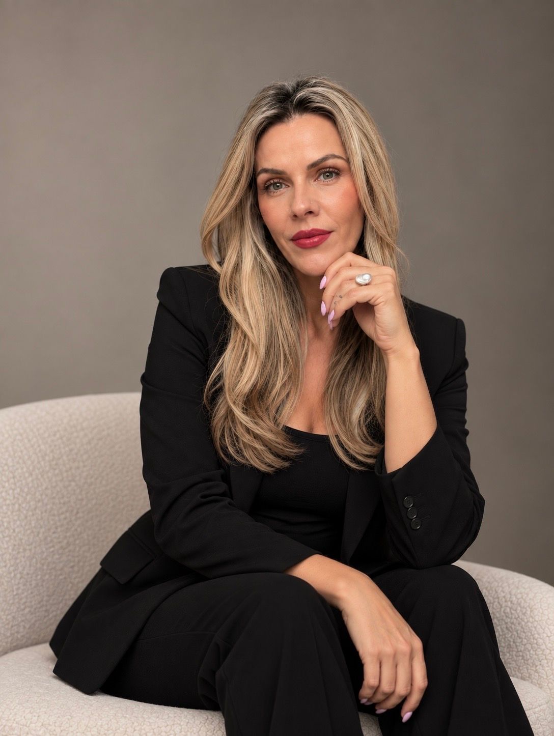 A confident woman in a black blazer sits in a white chair, with long blonde hair and red lips.
