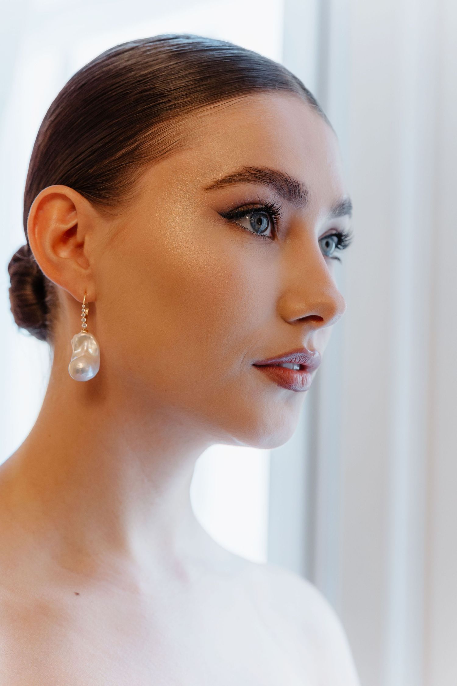 Close-up profile of bride with sleek updo wearing large baroque pearl drop earrings by bright window.