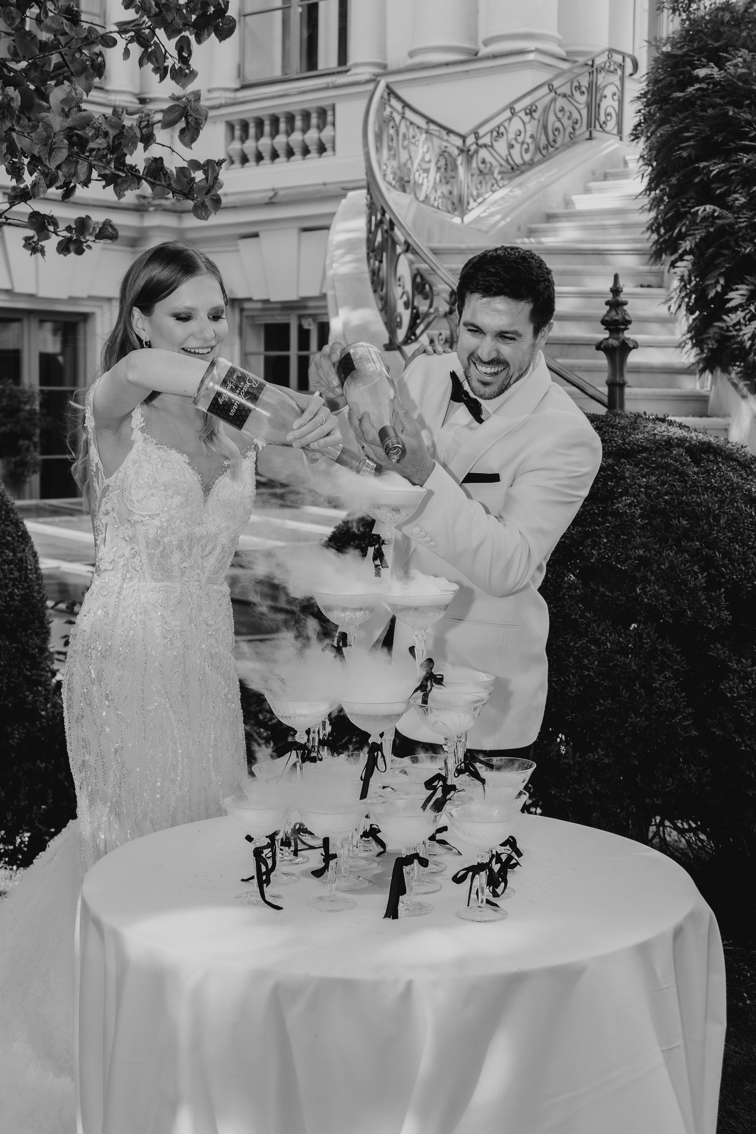 Bride and groom pouring champagne tower at outdoor wedding reception in black and white photo.