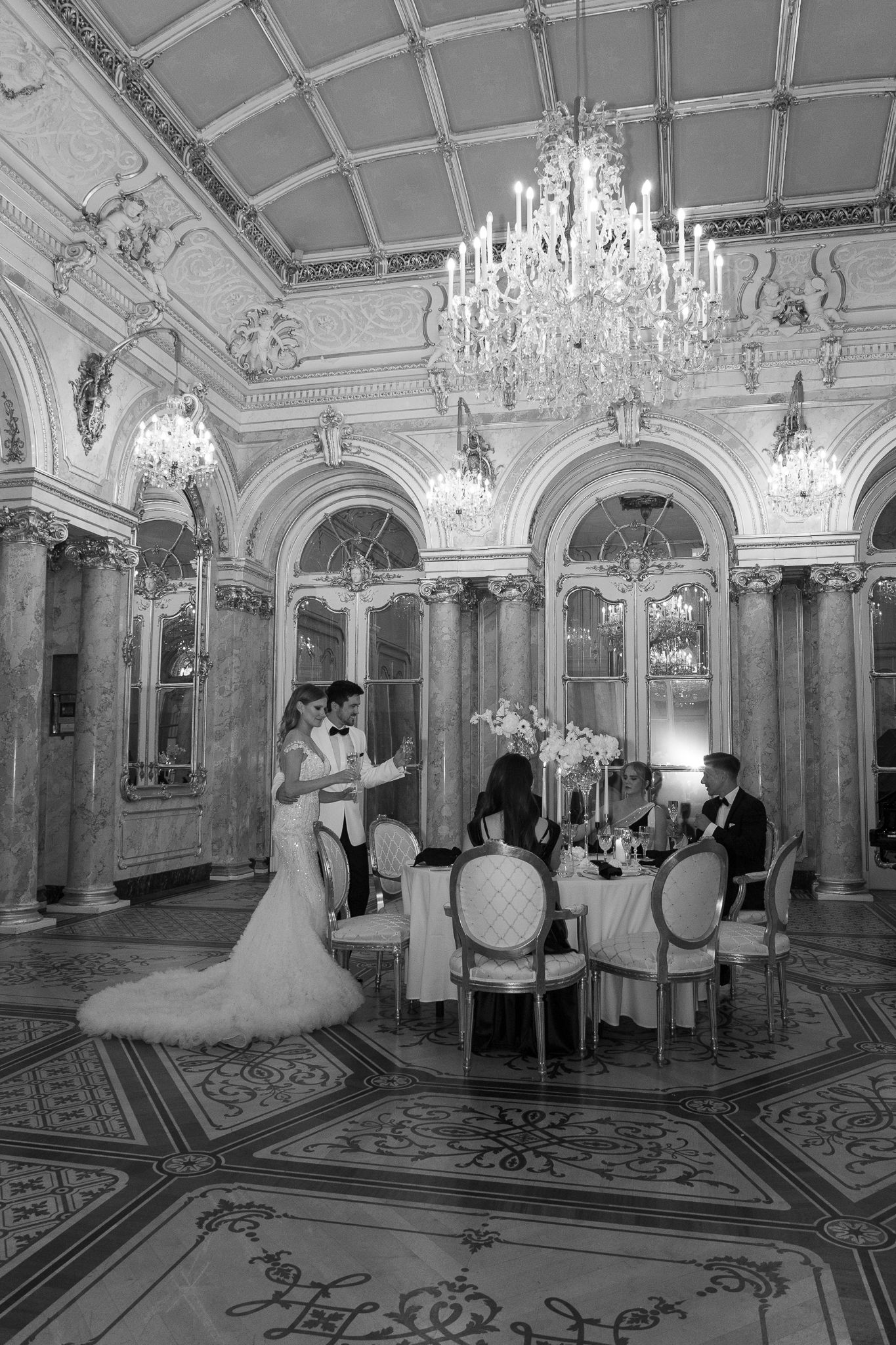 Bride and groom share first dance in opulent ballroom with crystal chandelier in black and white.