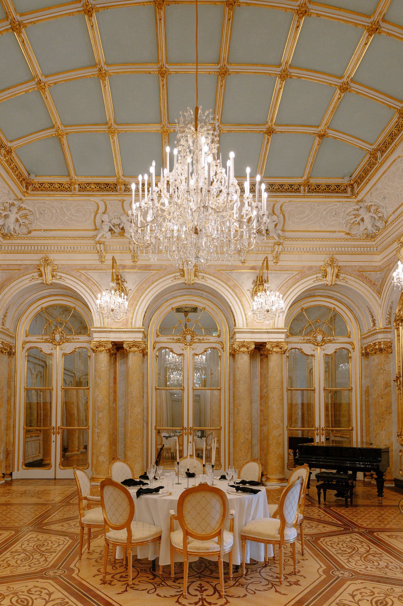 Opulent gold and white ballroom with crystal chandelier, marble columns, arched mirrors, and grand piano for wedding reception.