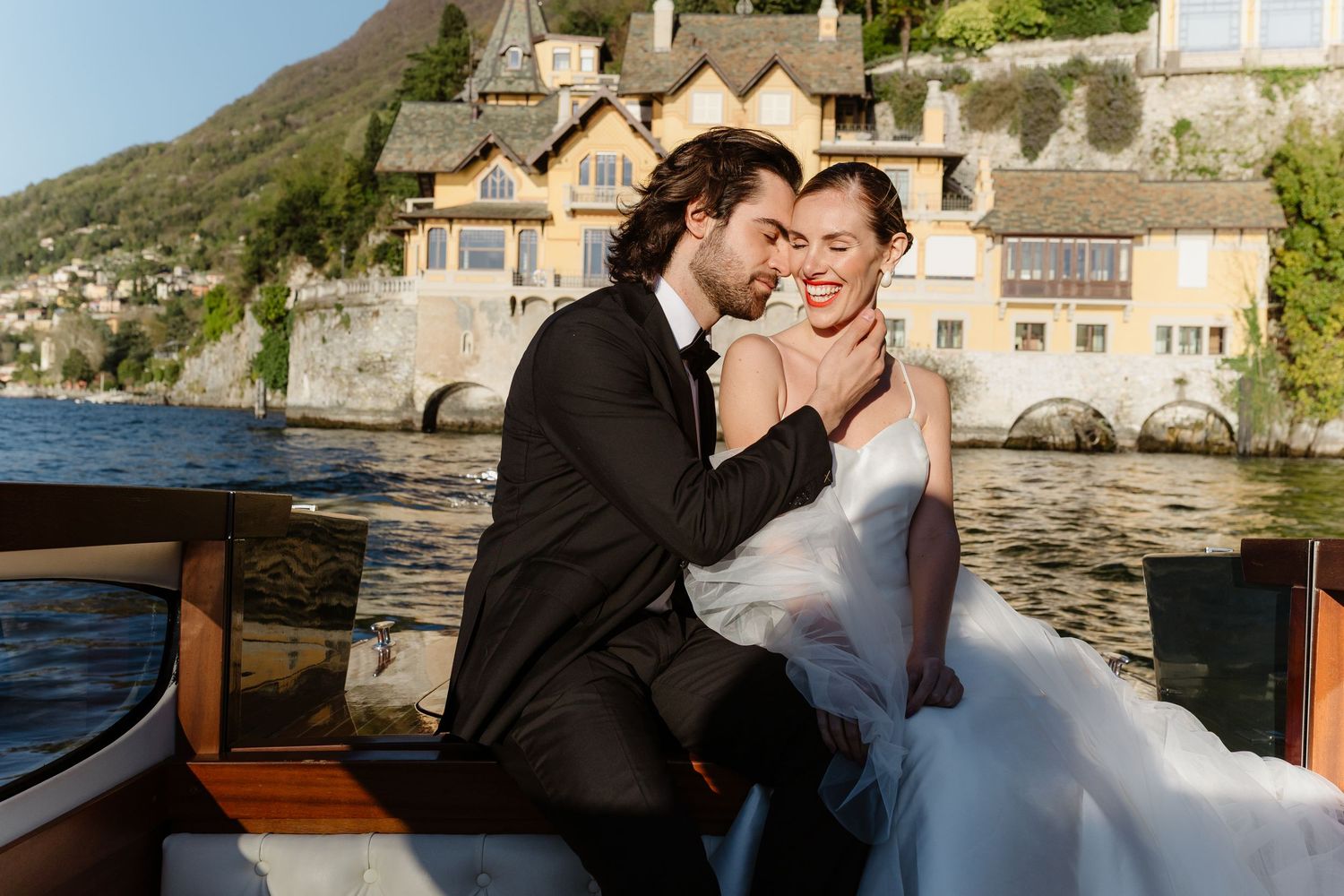Romantic couple embracing on a wooden boat with a golden lakeside villa and mountains behind them.