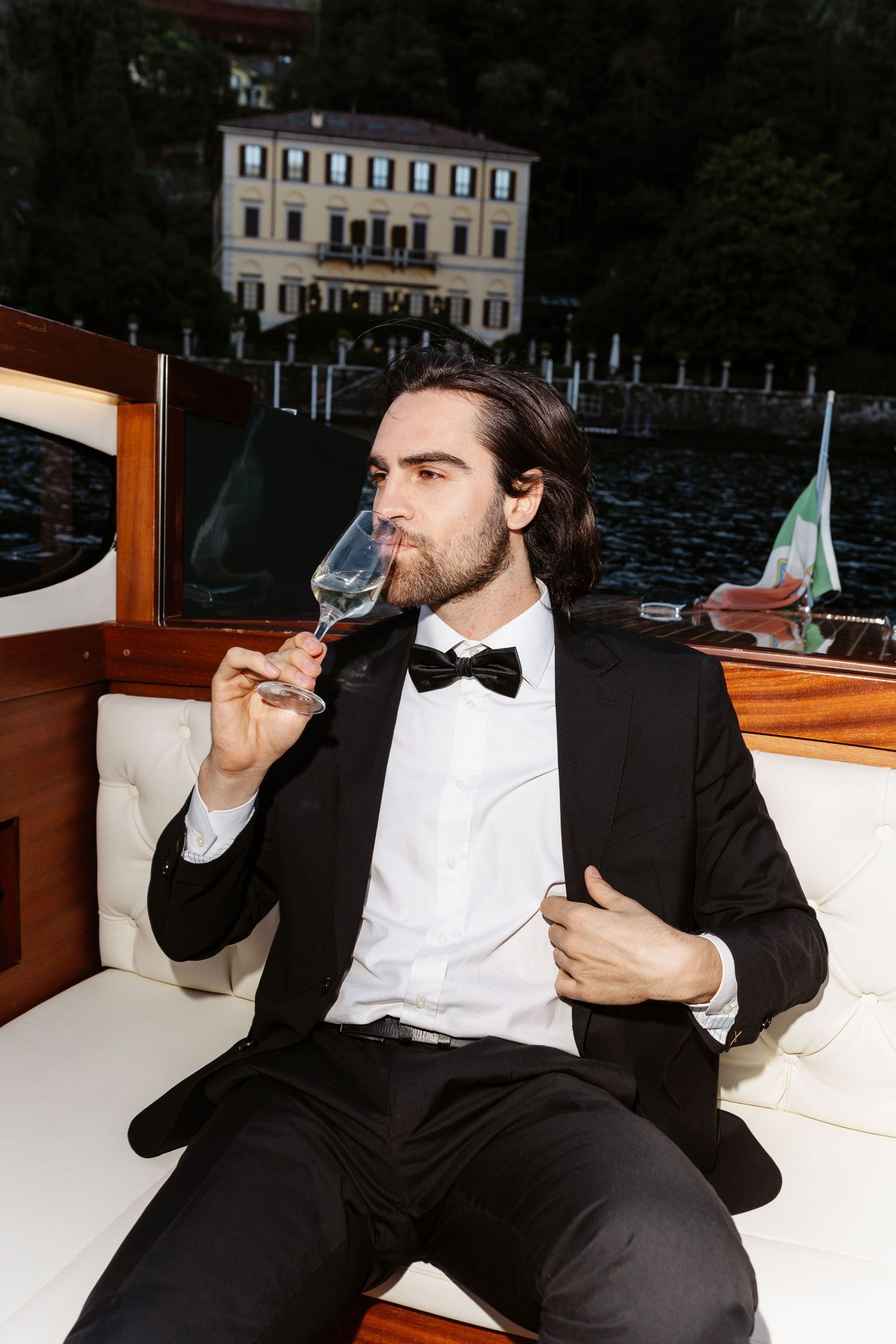 Dapper groom in black tuxedo drinking champagne on a wooden boat with Italian villa in background.