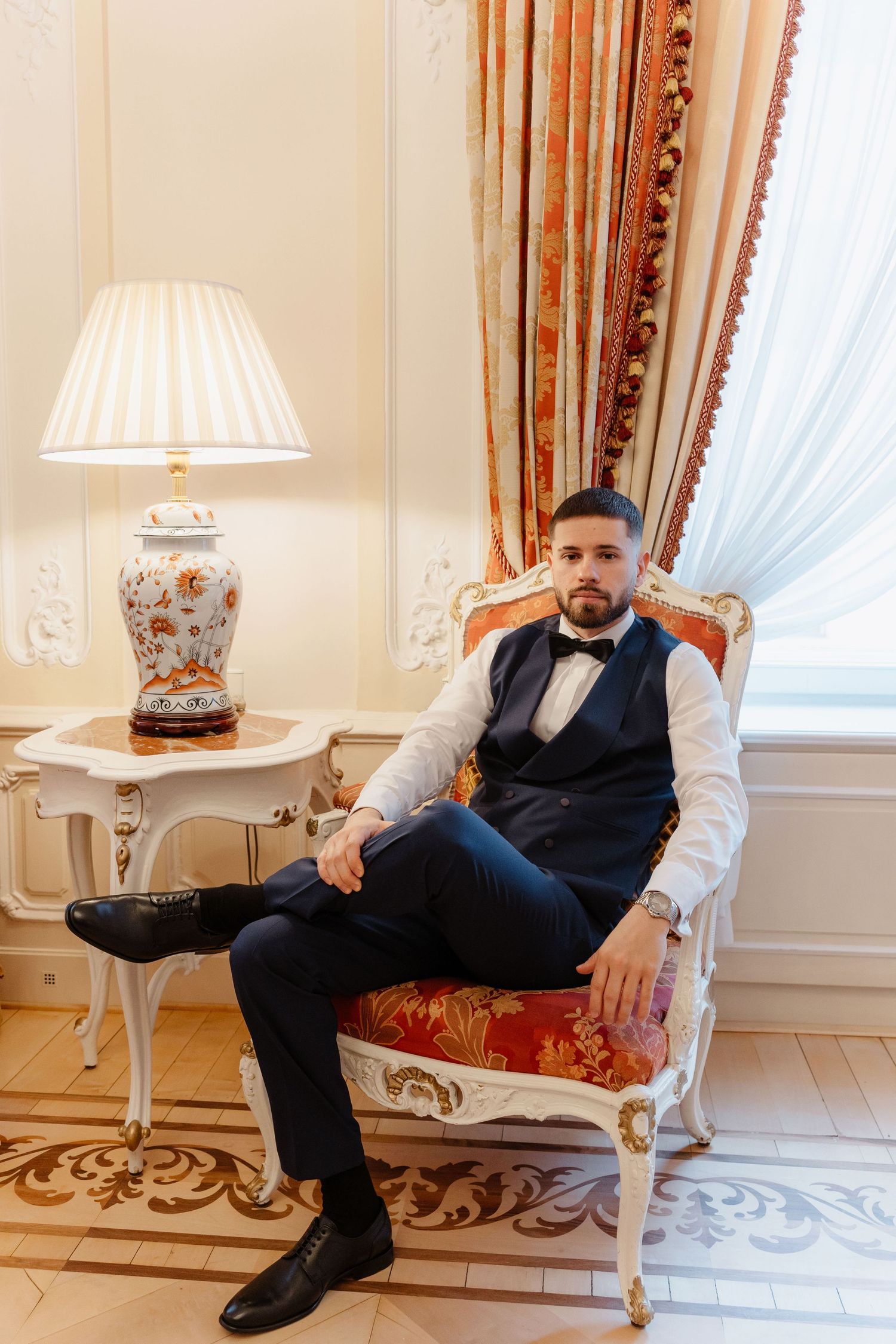 Dapper groom in navy blue suit with bow tie seated in ornate gold chair in luxurious room.