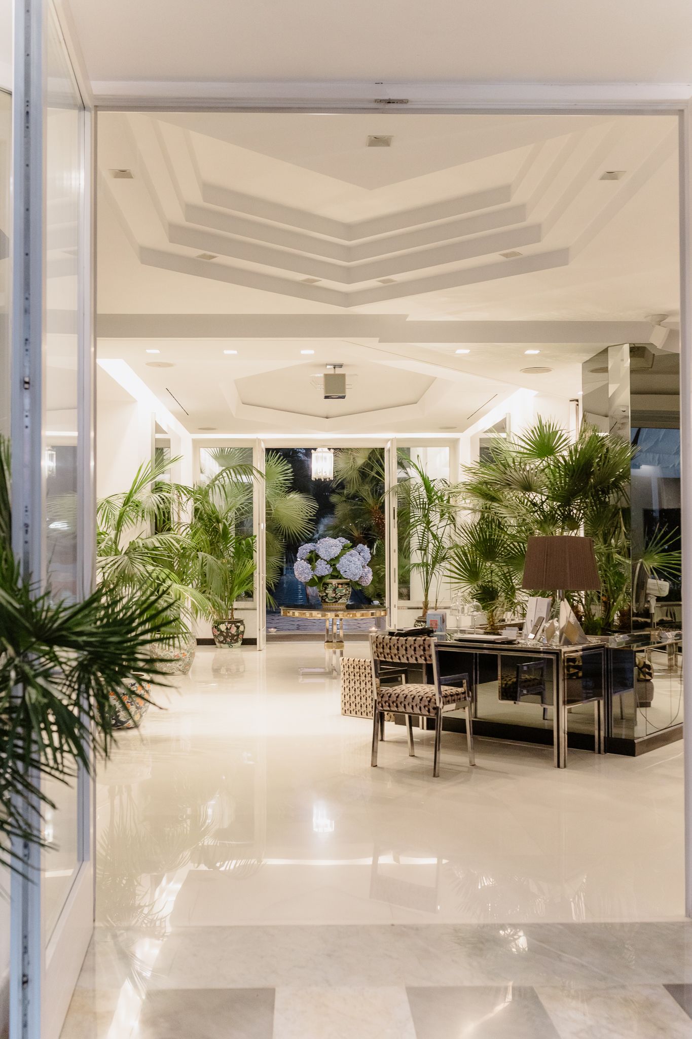Elegant hotel lobby with marble floors, tropical plants, mirrored furniture, and a decorative ceiling in a luxurious interior.
