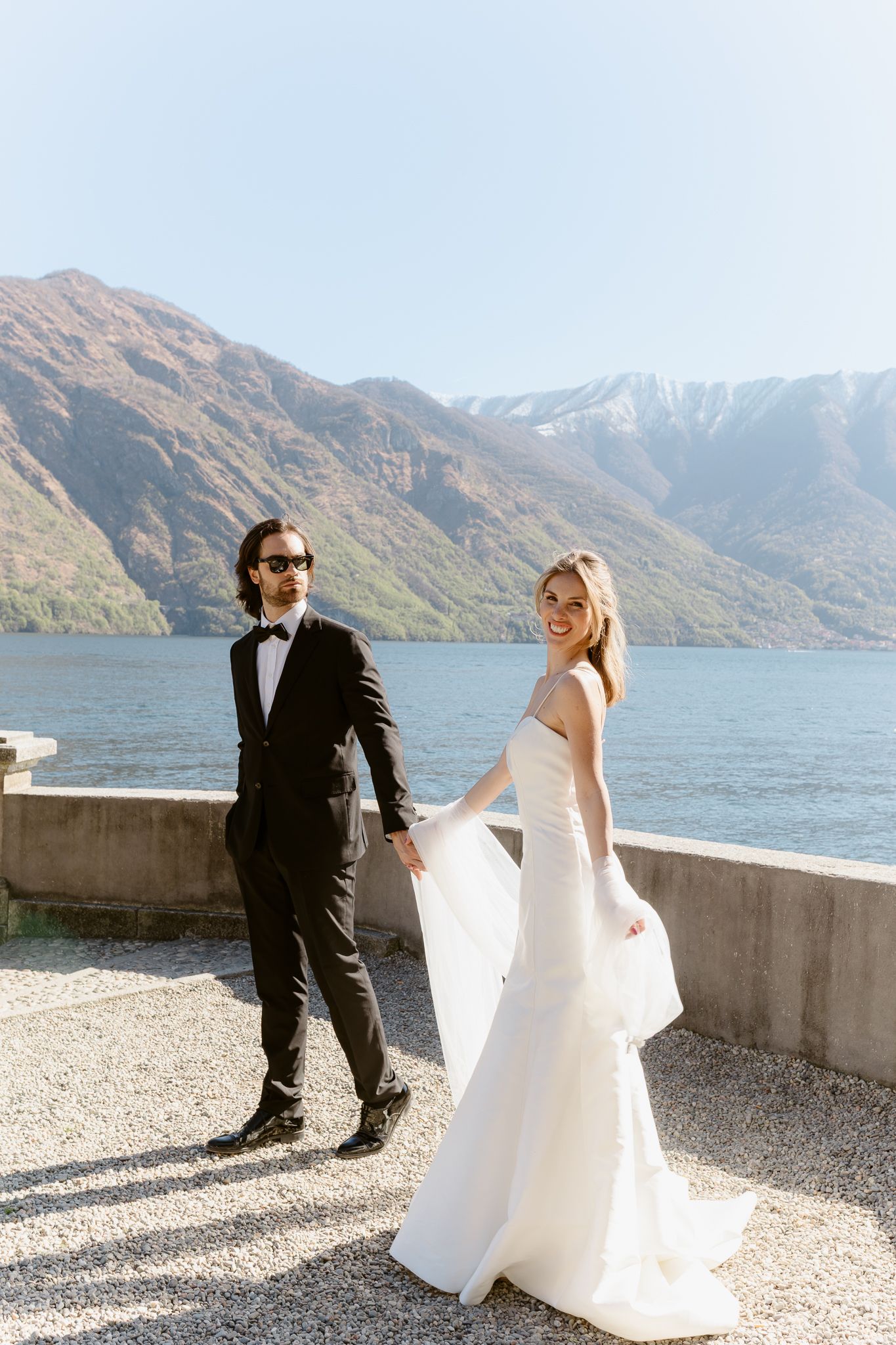 Smiling bride in white mermaid gown holds grooms hand while walking along scenic Lake Como waterfront.