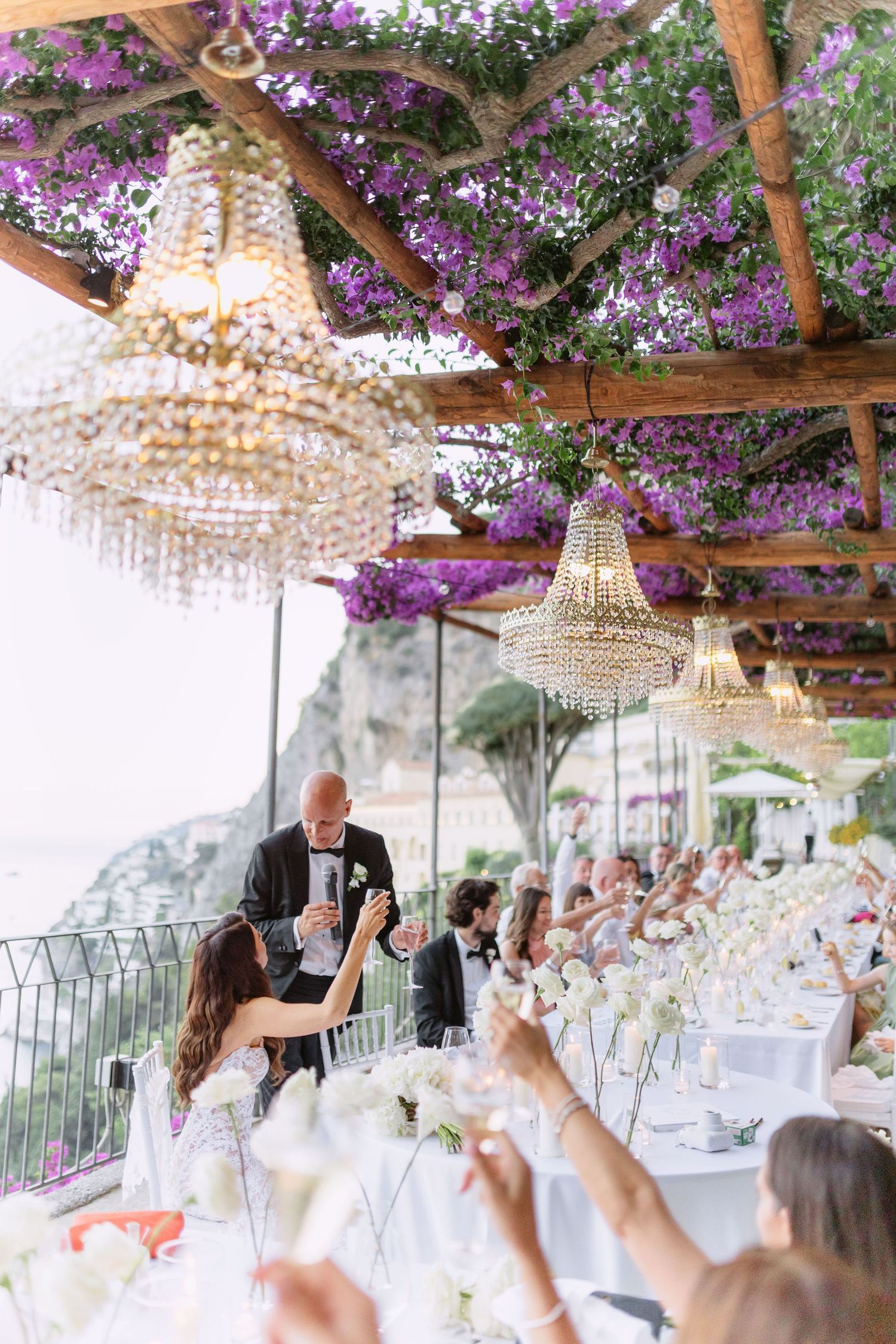 Elegant outdoor wedding reception under pergola with crystal chandeliers and purple bougainvillea, guests raising champagne glasses.
