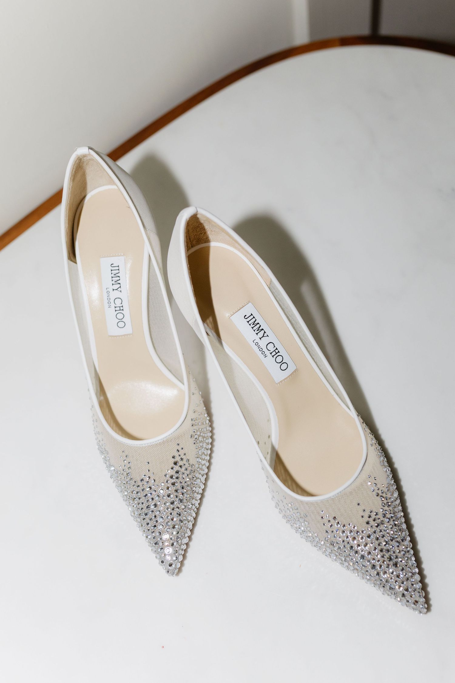 Jimmy Choo crystal-embellished white pointed-toe heels displayed on a marble surface.Editorial wedding photography.