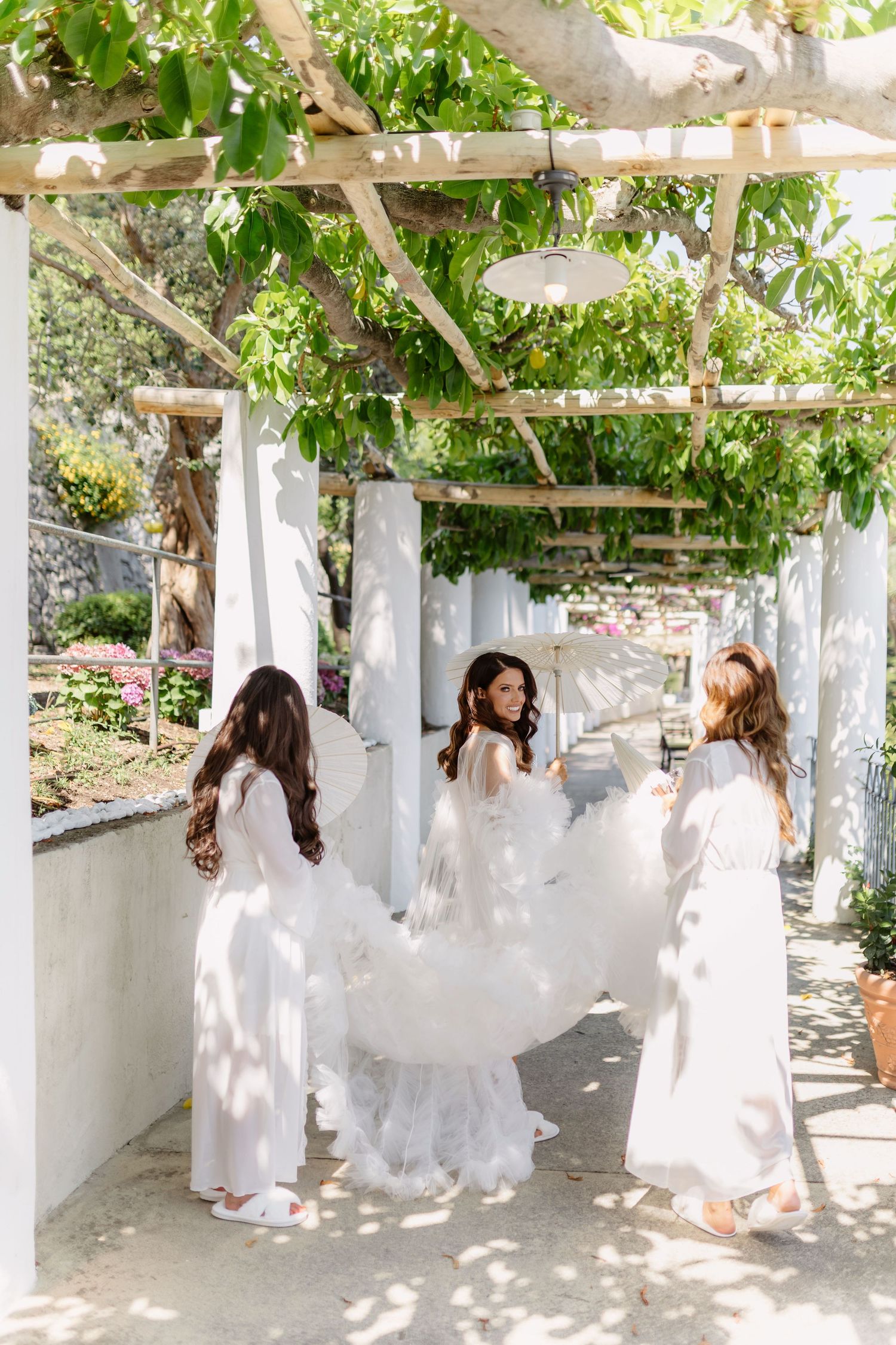Bridesmaids in white dresses help bride with flowing tulle gown under pergola with lush greenery at outdoor wedding.