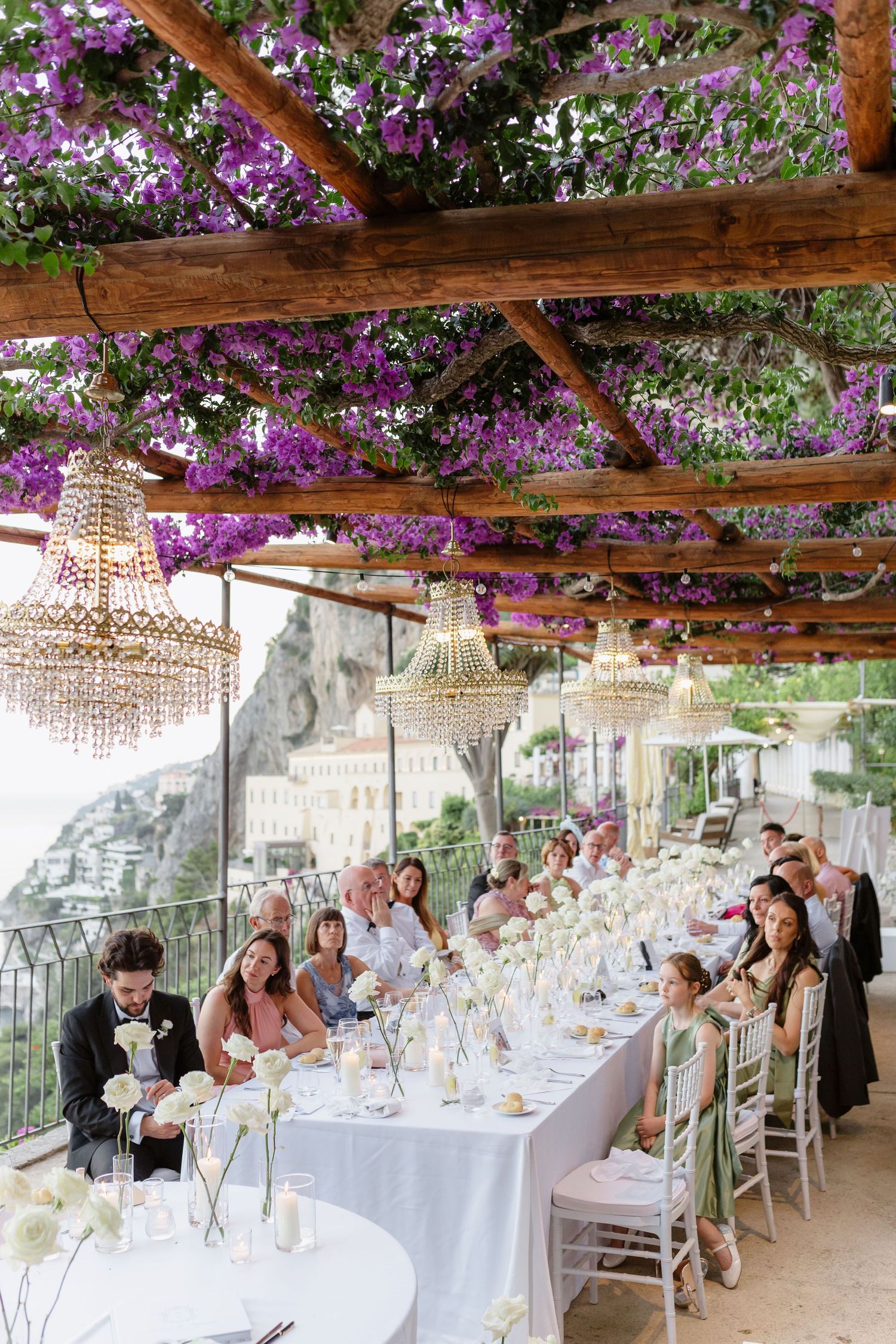 Elegant outdoor wedding reception under a pergola adorned with purple wisteria and crystal chandeliers on the Amalfi Coast.