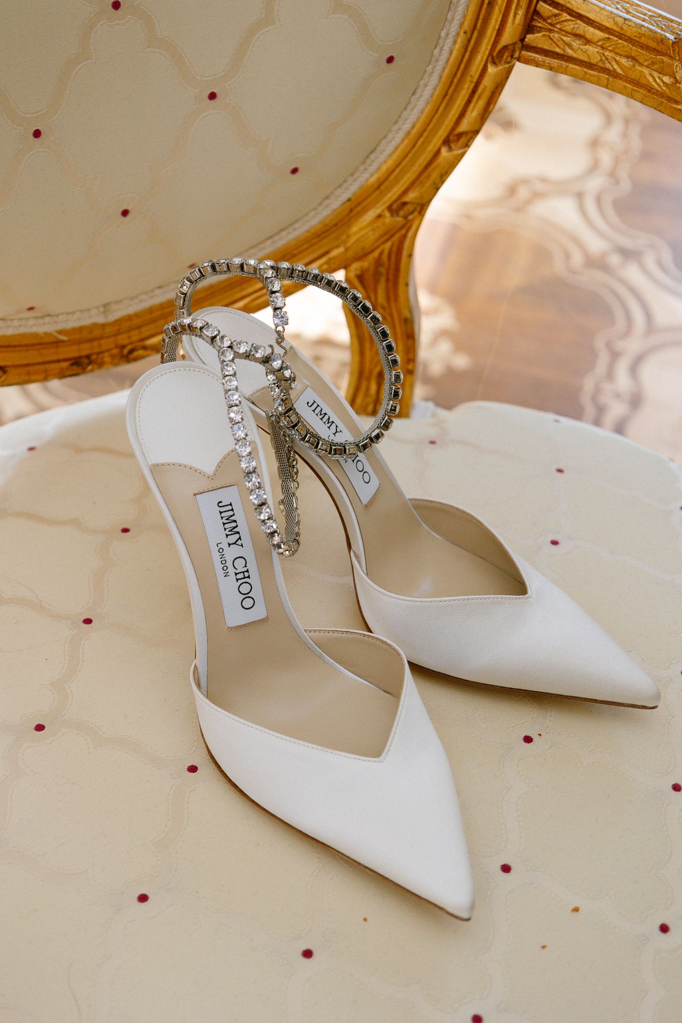 White Jimmy Choo bridal heels with crystal ankle straps displayed on ornate gold-framed chair.