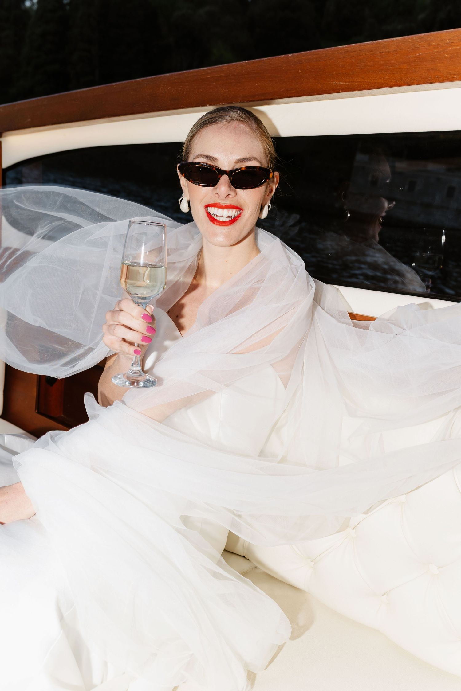 Smiling bride in white wedding dress and sunglasses holding champagne flute while seated on a luxury boat.