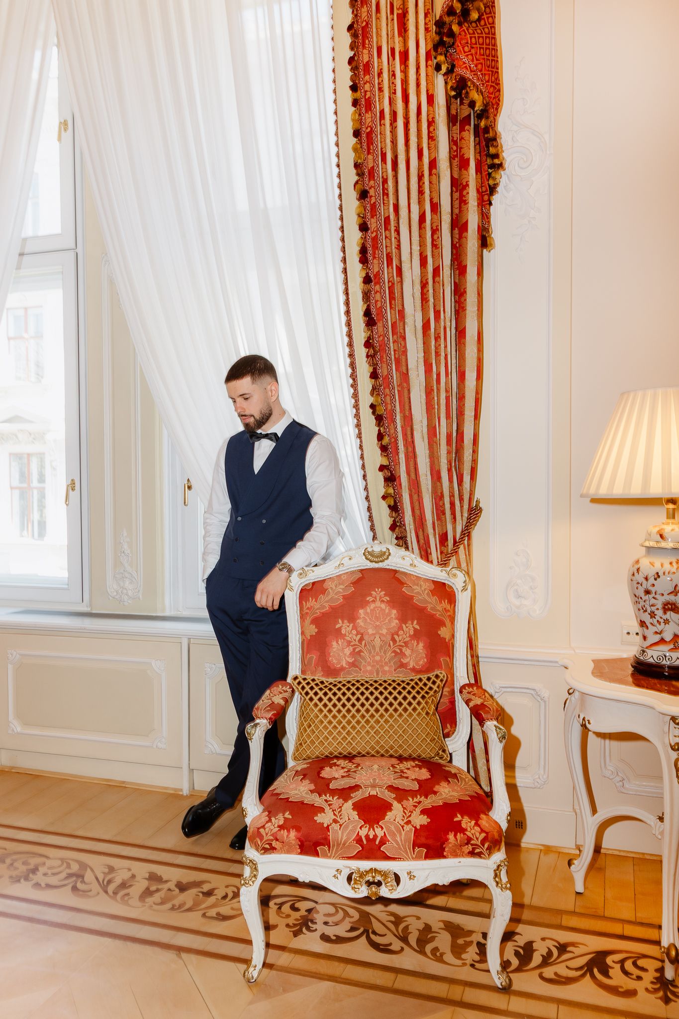 Dapper groom in navy blue vest and bow tie stands beside ornate red gilded chair near draped window.