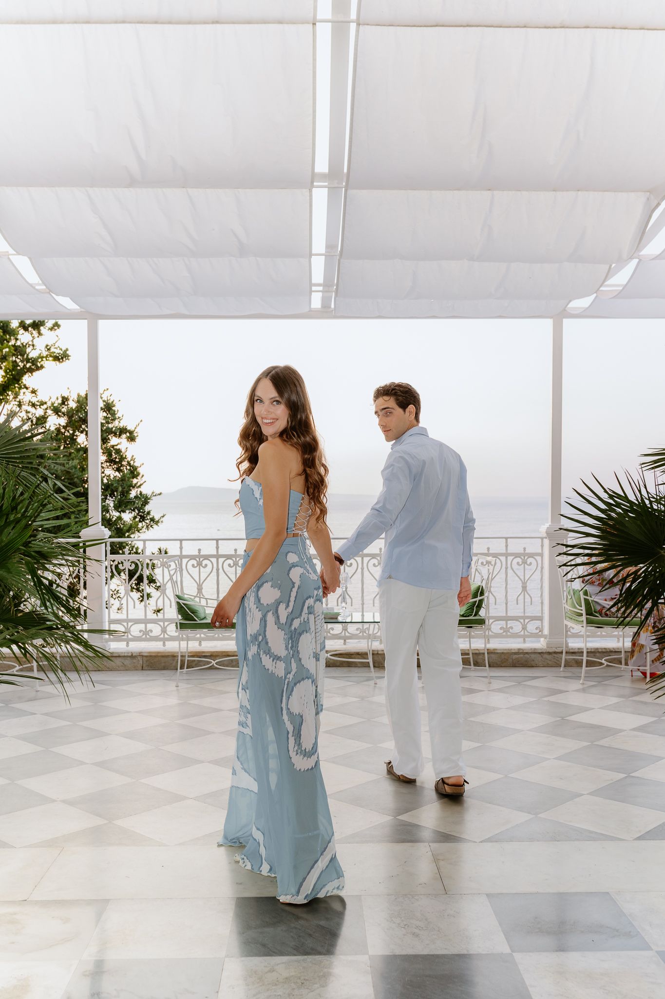 Elegant couple holding hands on a marble terrace, woman in blue floral gown with ocean view backdrop.