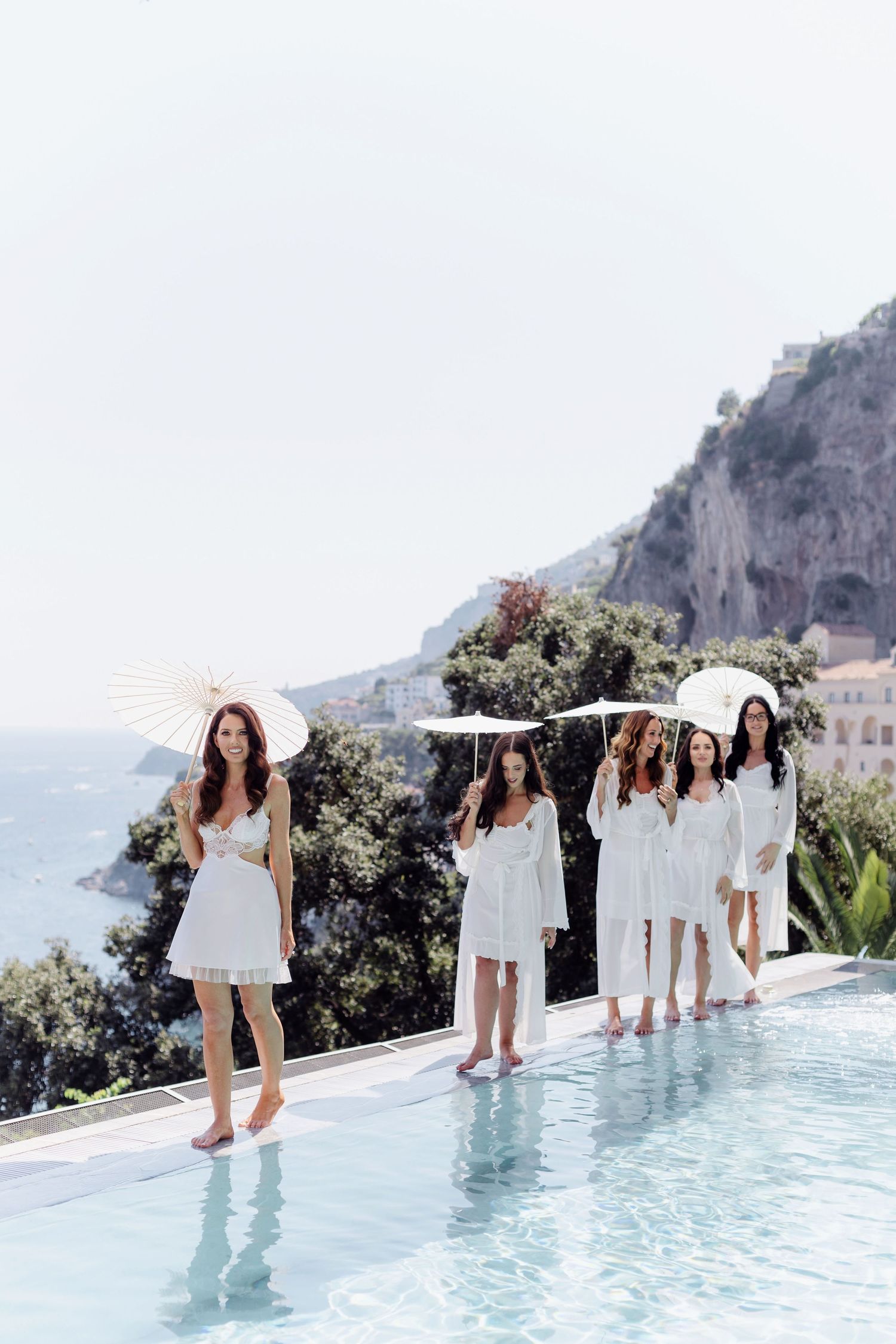 Five women in white dresses holding parasols walk along an infinity pool with scenic Amalfi Coast cliffs in the background.