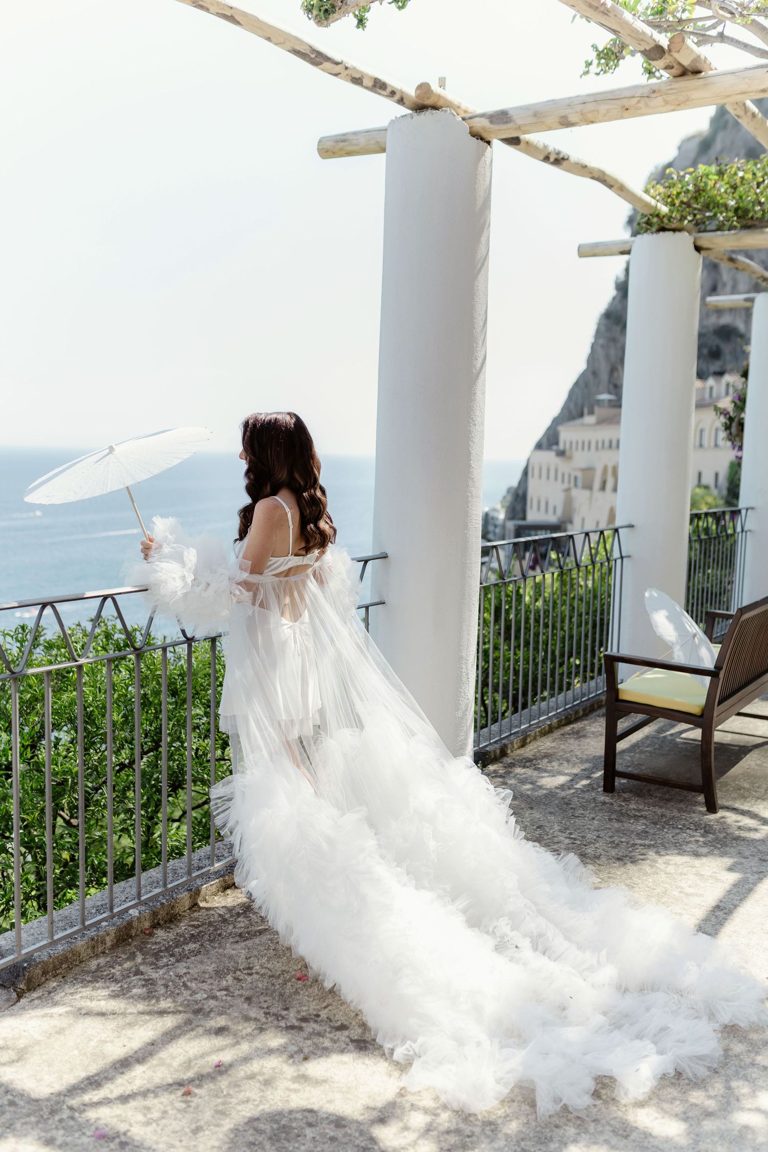 Bride in white tulle gown holding parasol on scenic Amalfi Coast terrace overlooking the sea.