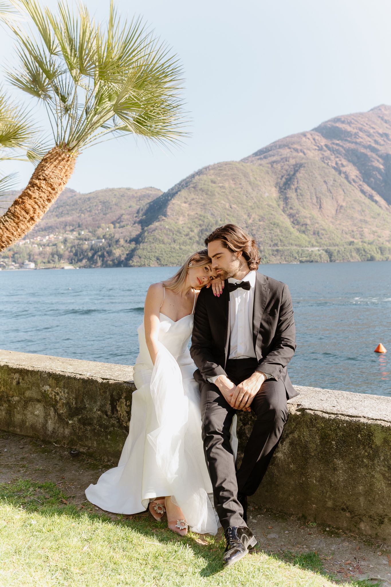 Newlyweds sit closely together on a stone bench near a palm tree overlooking beautiful Lake Como.