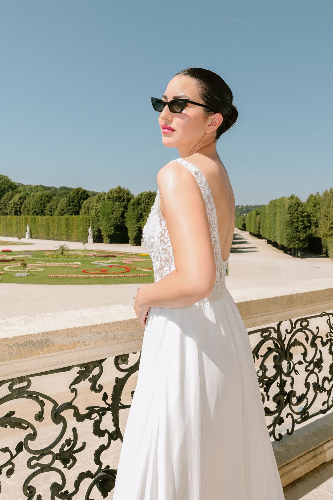 Bride in backless white gown and sunglasses poses on decorative iron balcony at a manicured formal garden in Schonbrün.