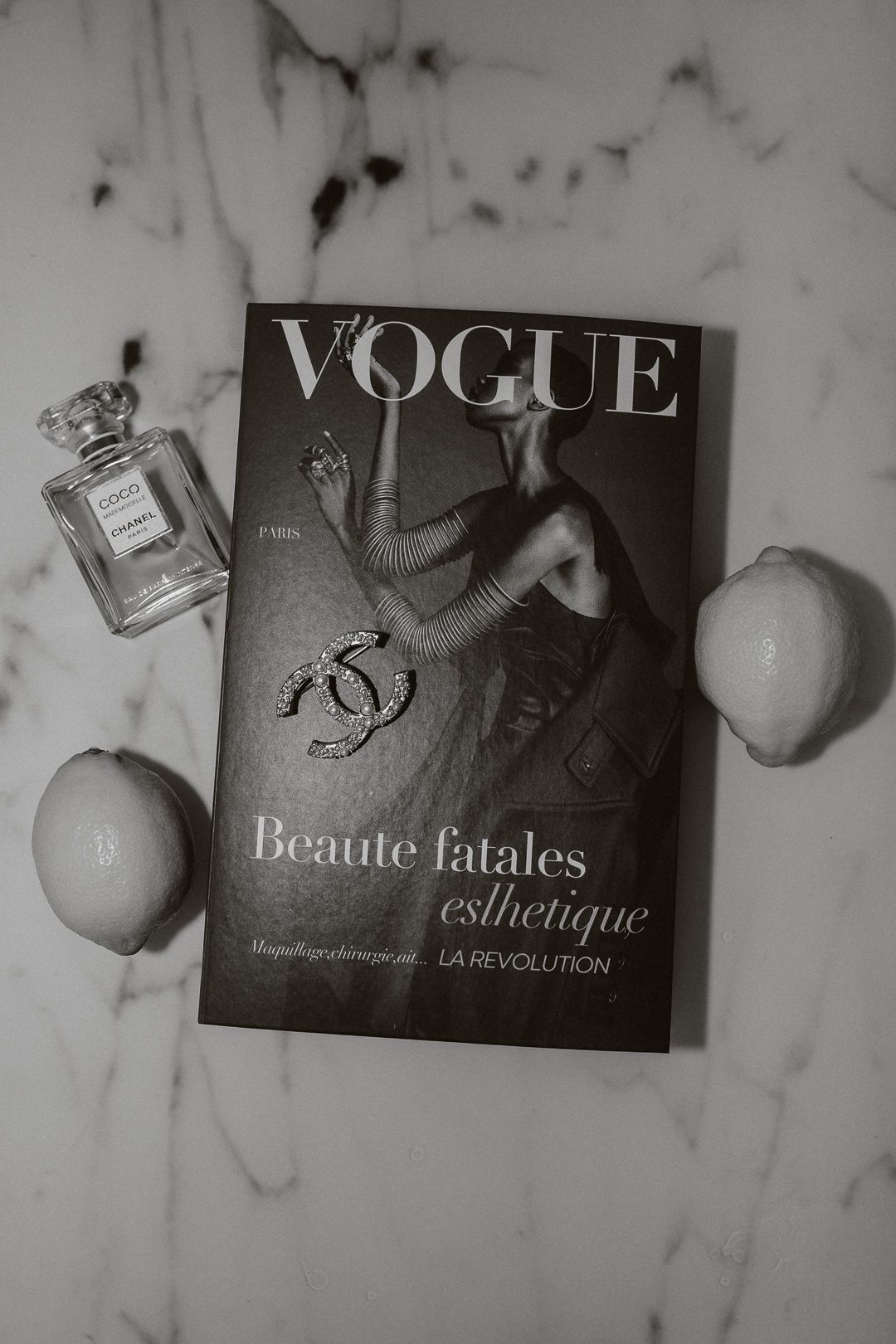 Vogue magazine with Chanel brooch, Coco Chanel perfume, and lemons on marble surface.