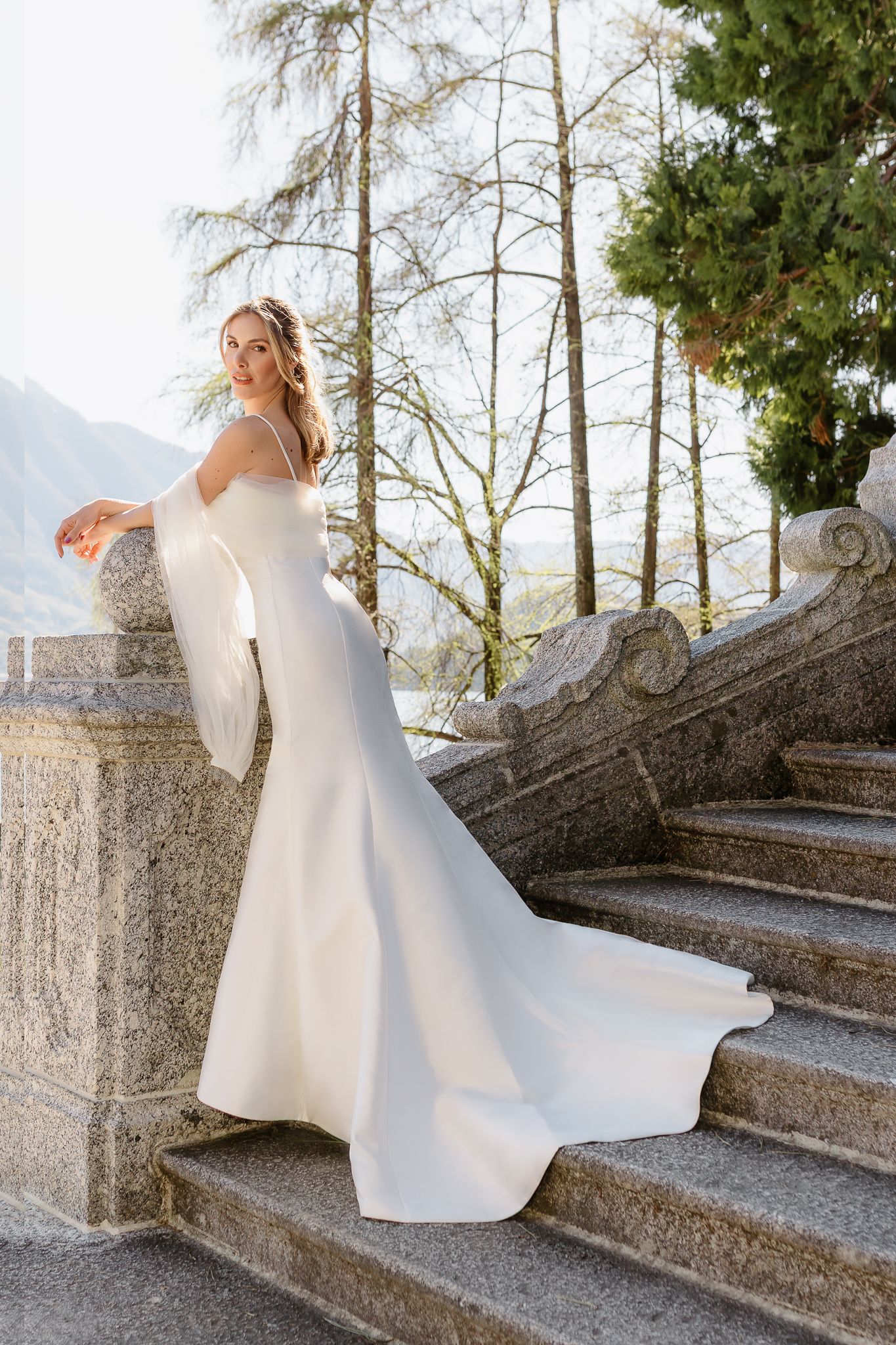 Elegant bride in white mermaid gown with sheer sleeves posing on stone steps at a lakeside villa.
