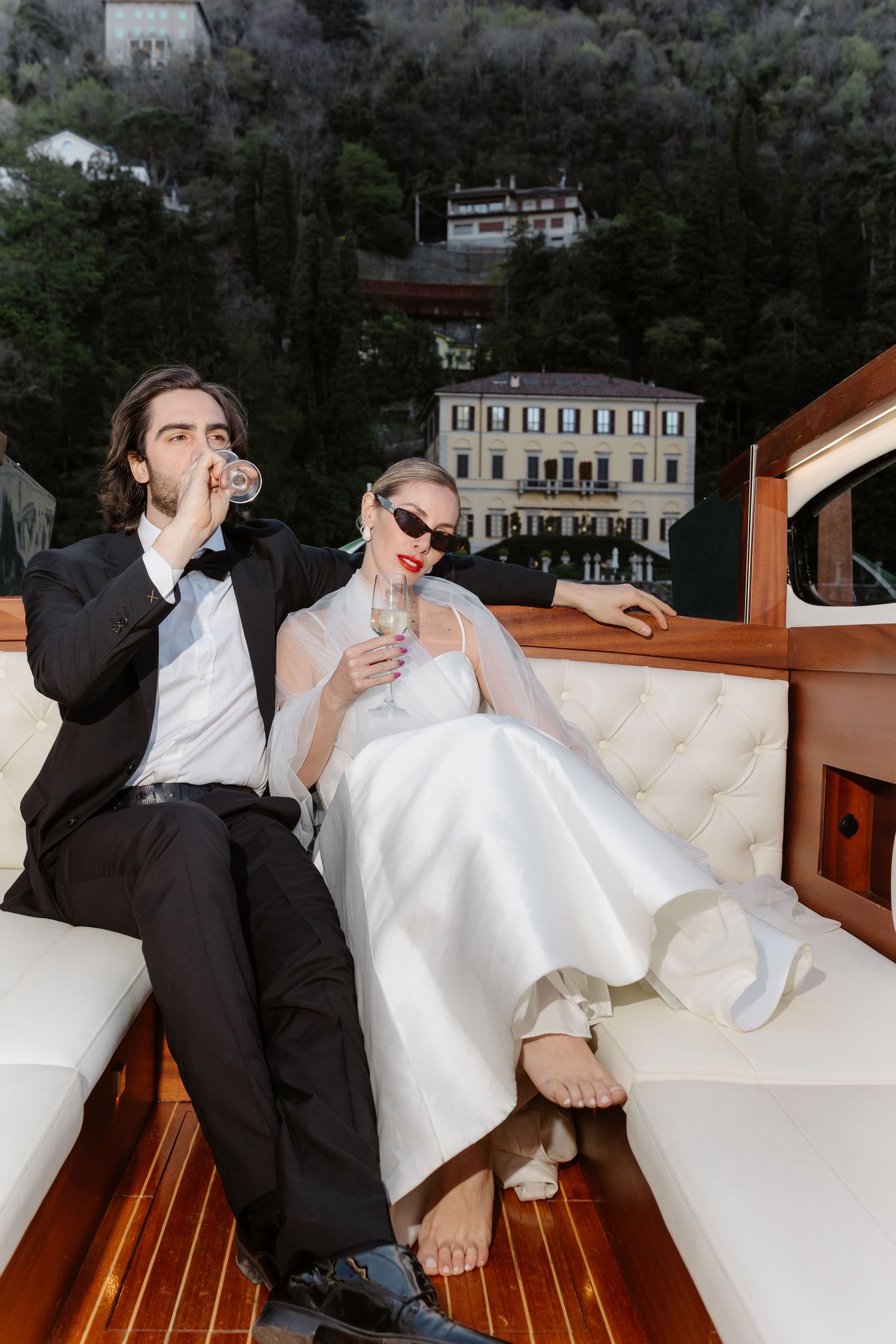 Bride and groom in wedding attire relaxing on luxury boat deck with champagne, Italian lakeside villa behind them.