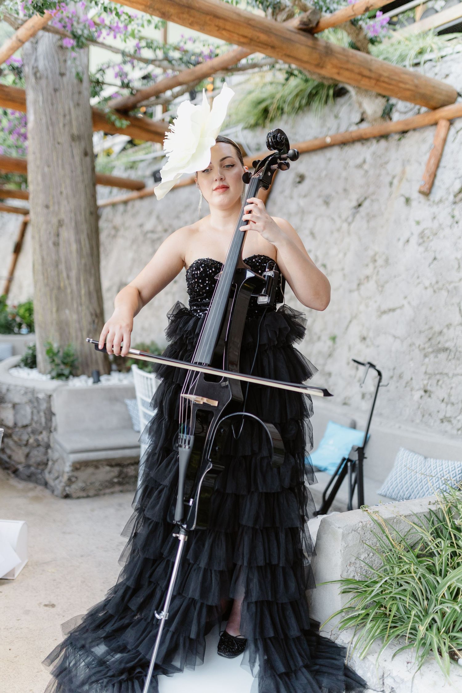 Woman in black ruffled gown playing cello outdoors, wearing white floral headpiece at elegant garden venue.