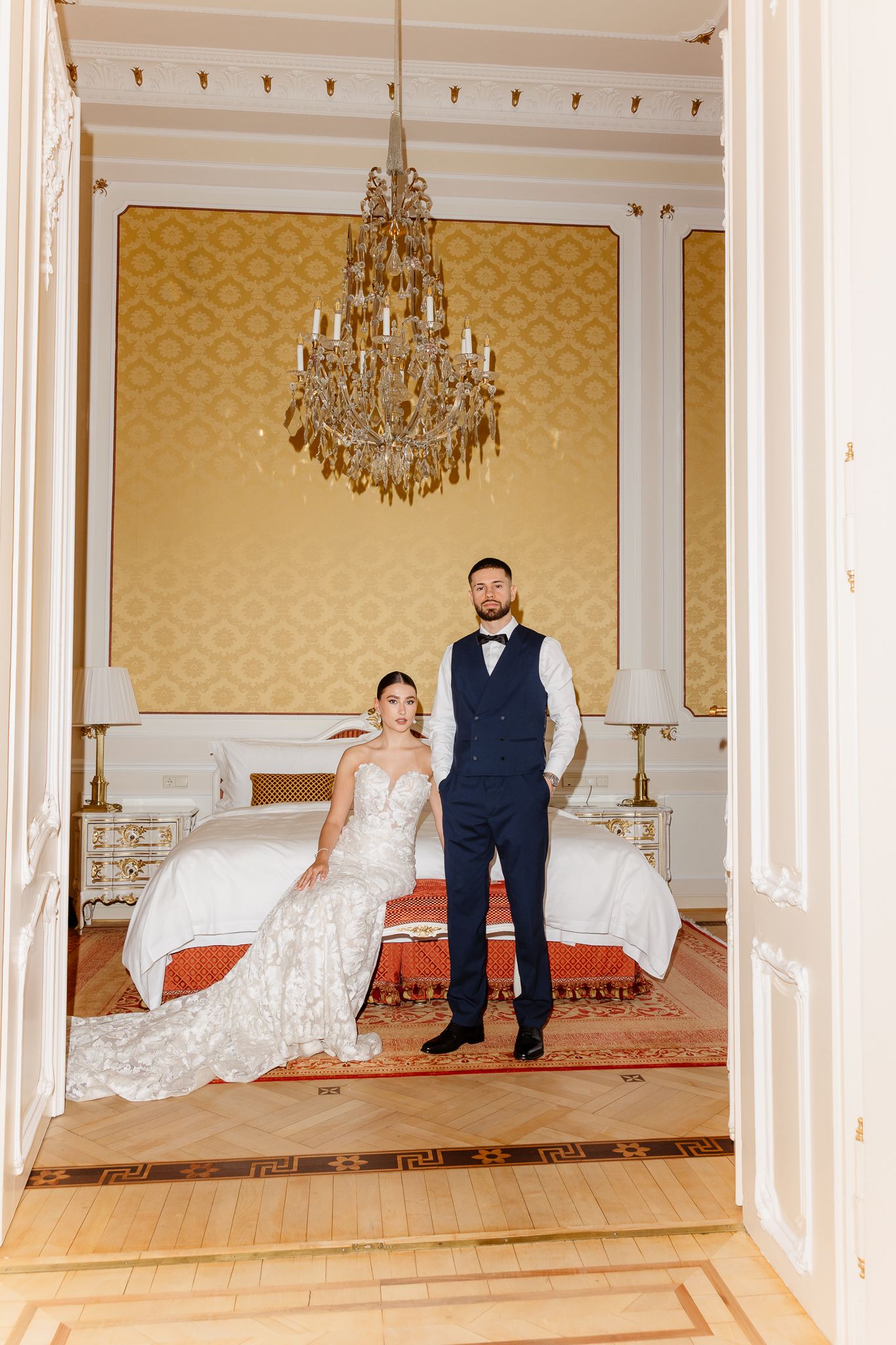 Bride and groom pose together in opulent gold wallpapered hotel suite beneath an elegant crystal chandelier.