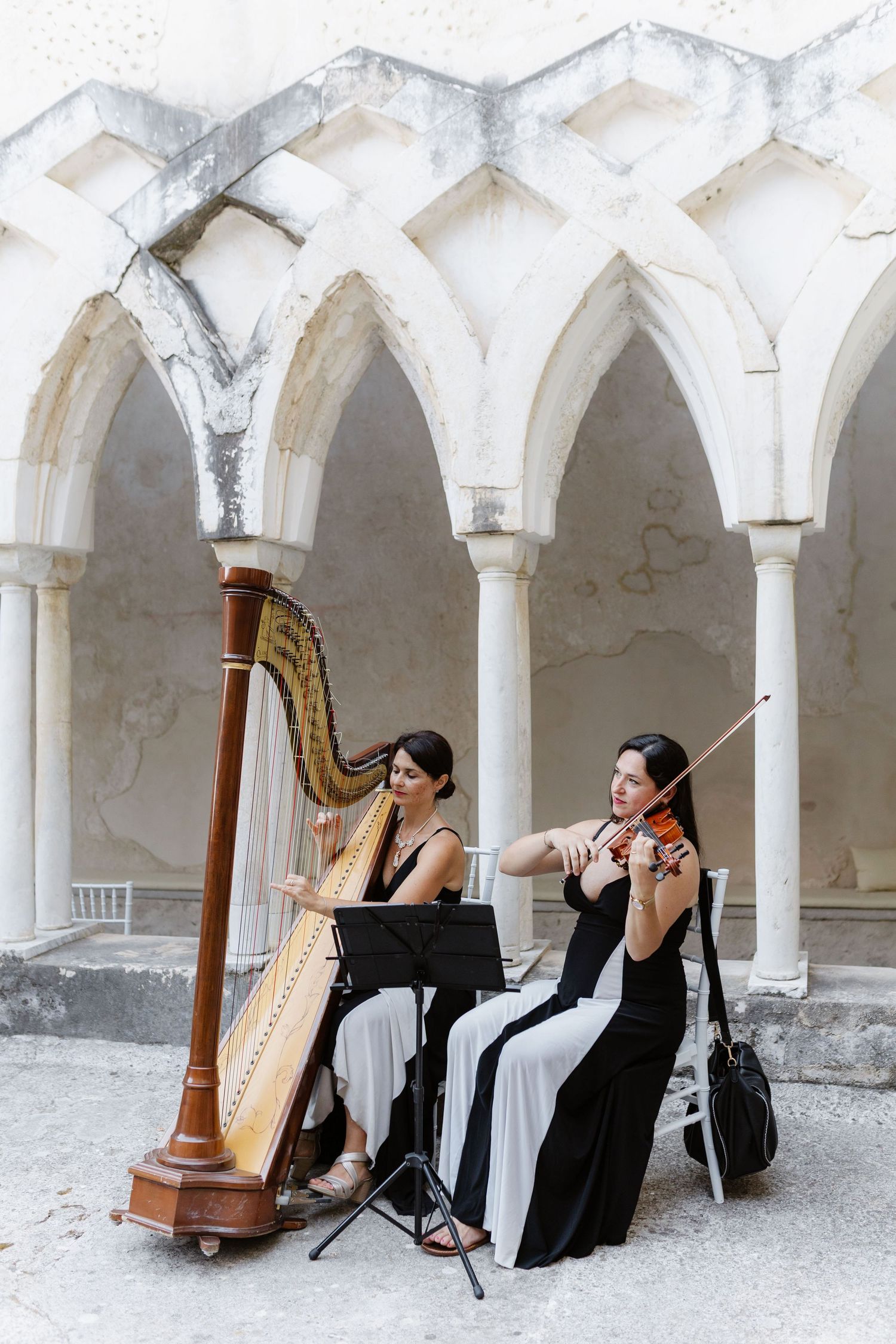 Two women in elegant gowns perform classical music on harp and violin in a white stone arched courtyard.