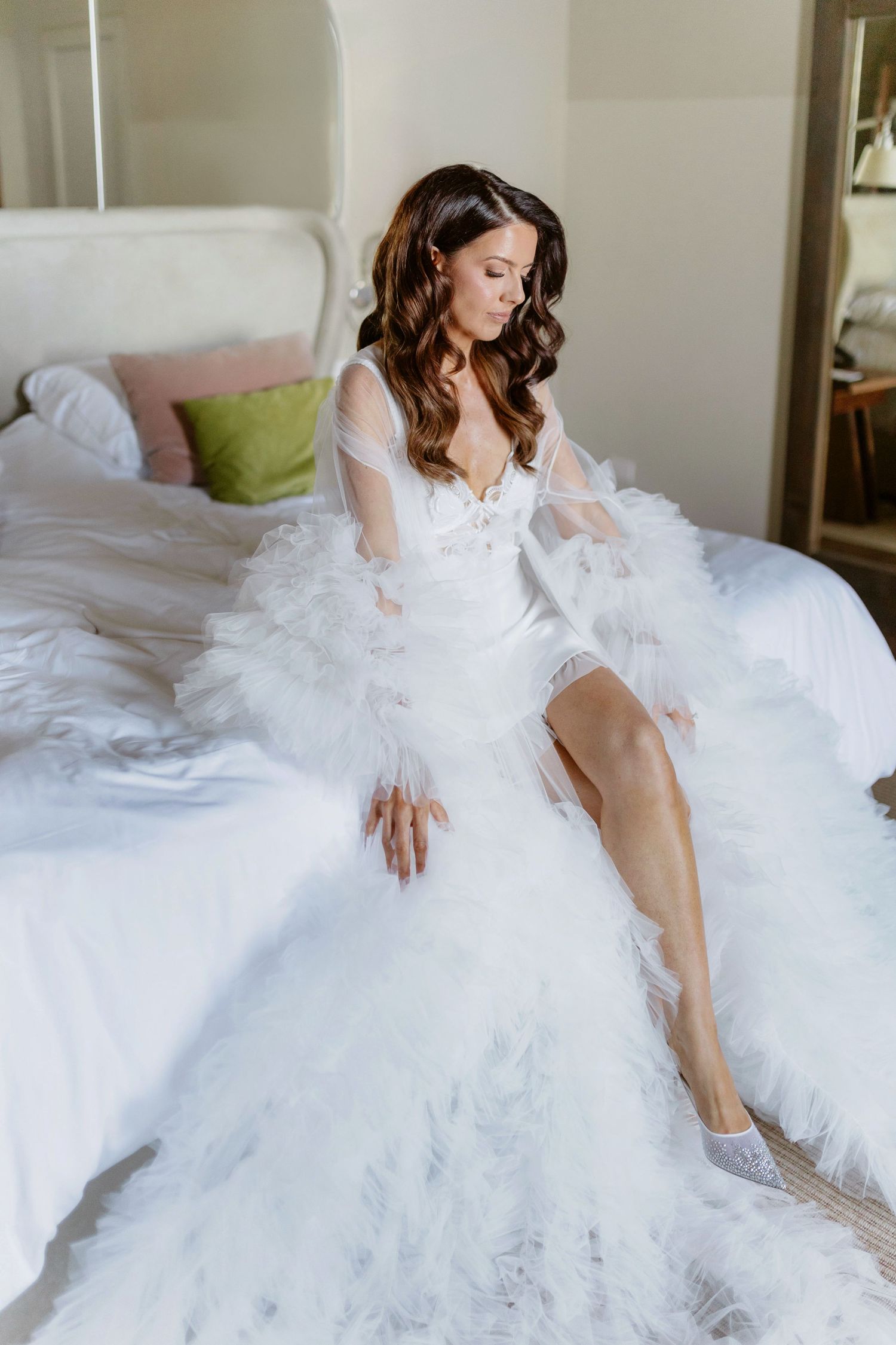 Bride in white feathered wedding gown with sheer sleeves sitting elegantly on a bed in a bright room.