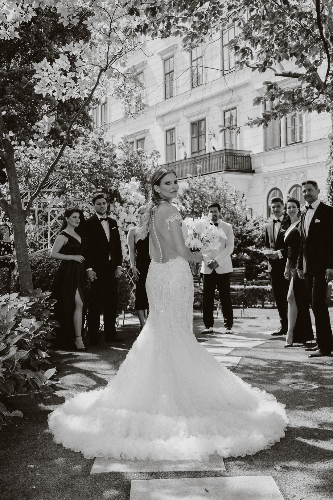 Bride in elegant mermaid gown with lace back and floral train walks toward groom at outdoor wedding ceremony.