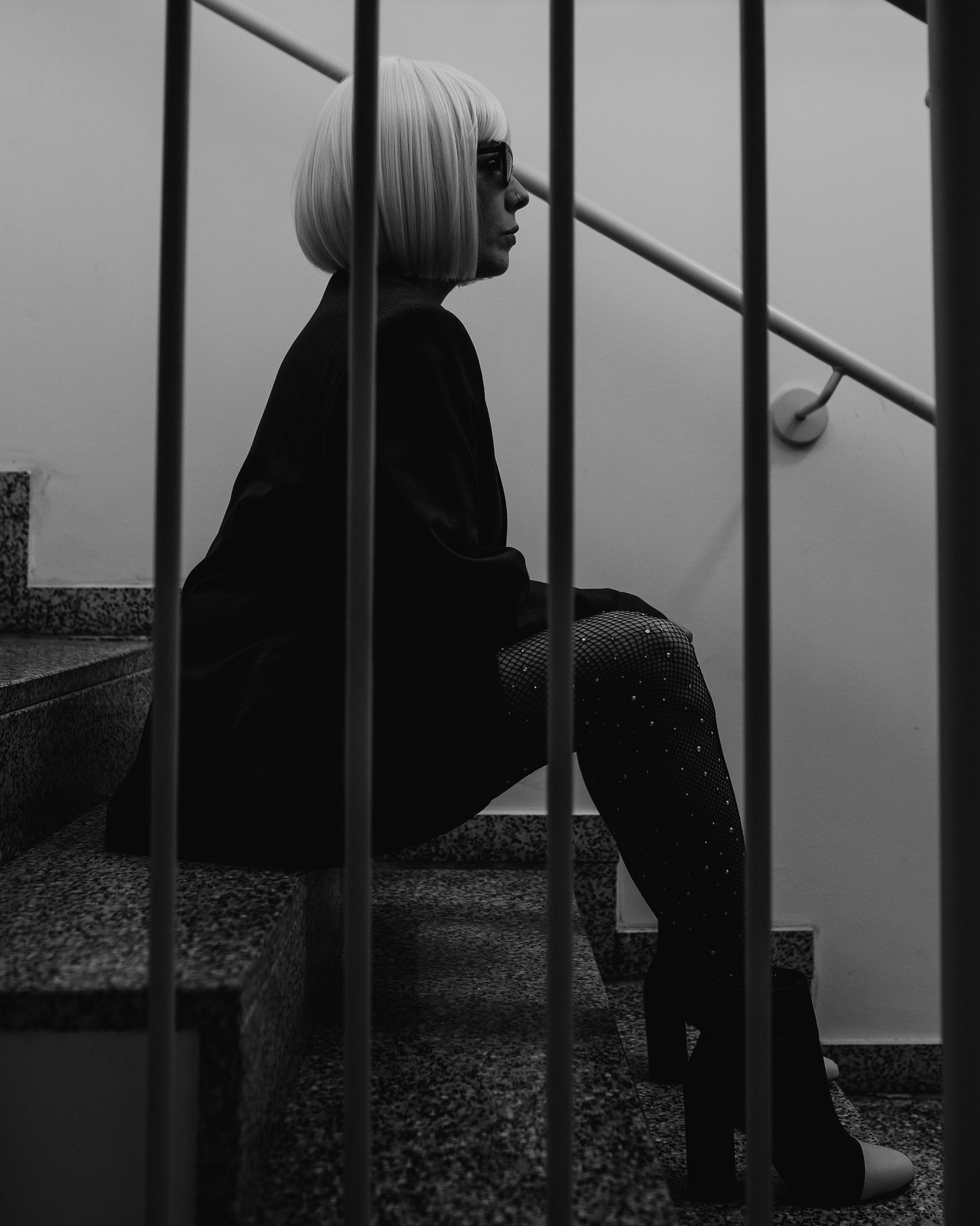 Black and white portrait of blonde woman in black outfit sitting on stairs behind bars.