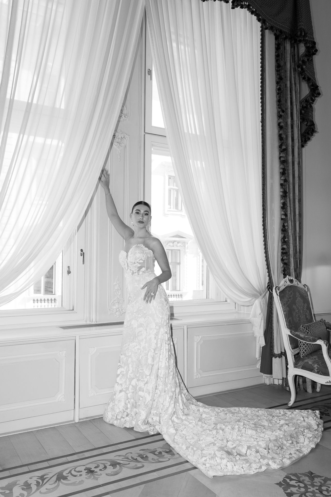 Black and white portrait of bride in floral lace mermaid gown holding sheer curtains by tall elegant window.