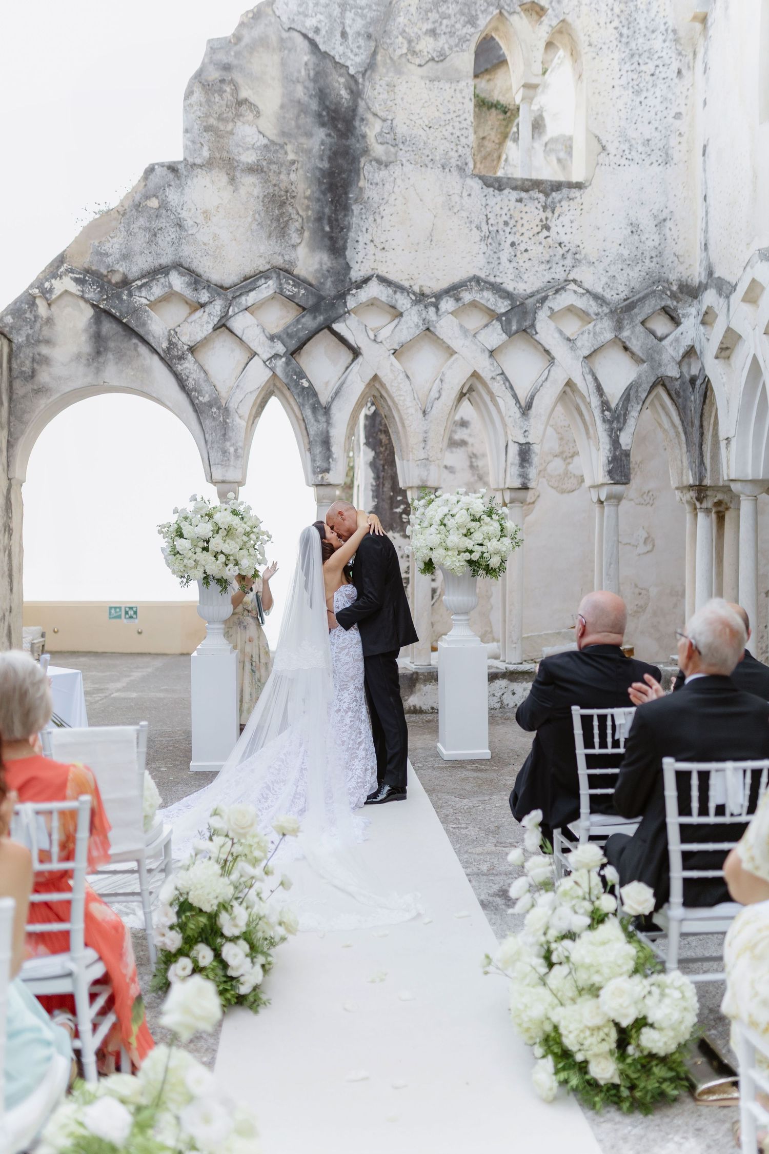 Bride and groom share first kiss at outdoor wedding ceremony in romantic Italian ruins with white floral arrangements in Anantara Convento