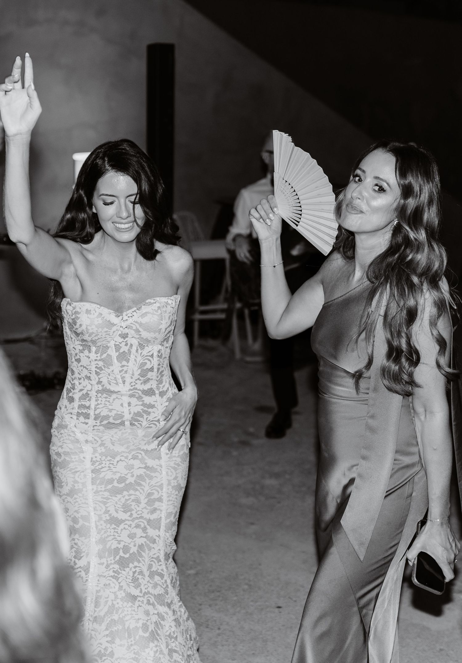 Bride in lace wedding gown dancing with bridesmaid holding fan at elegant wedding reception.