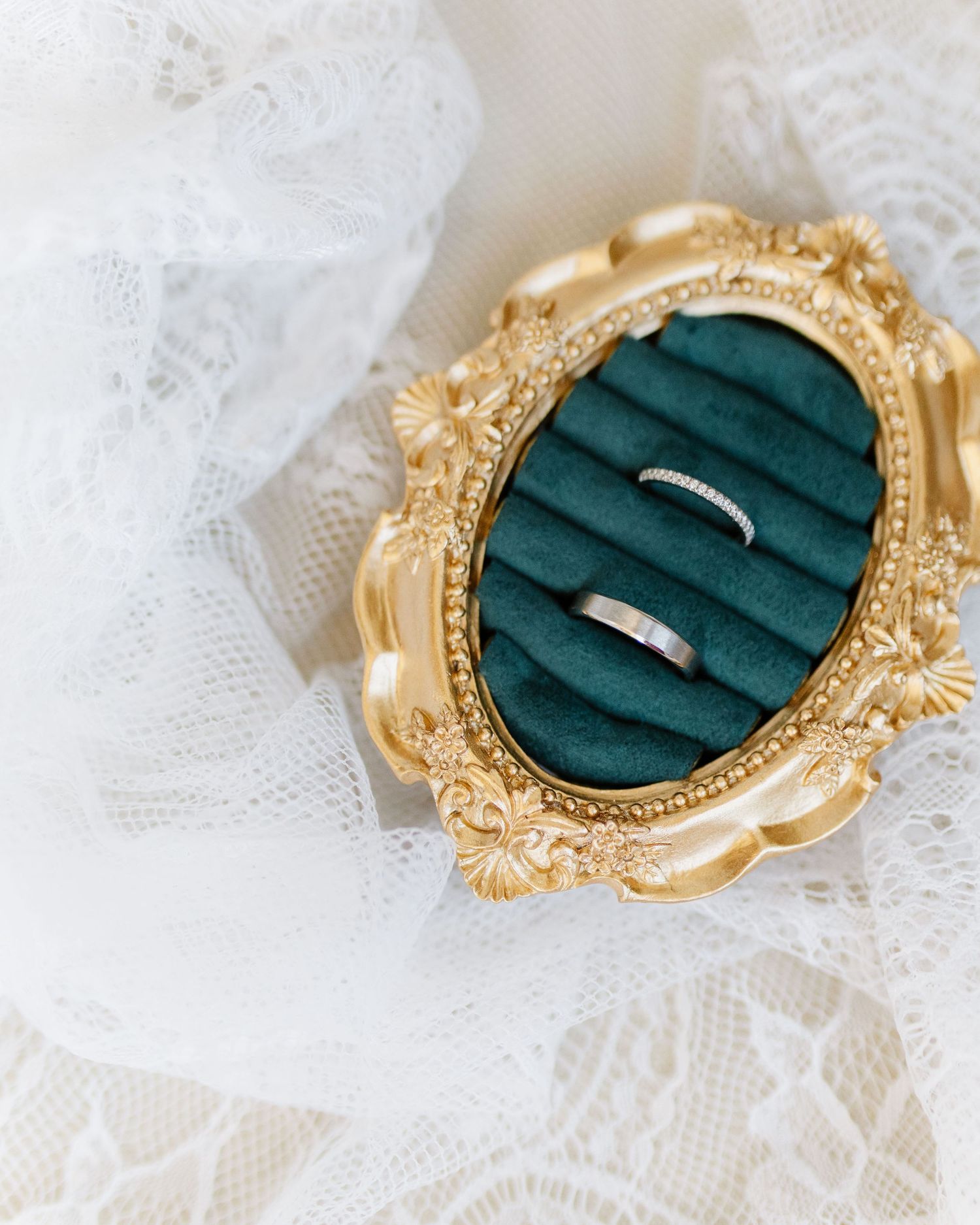 Two wedding bands displayed in an ornate gold oval ring box with green velvet lining, placed on white lace fabric.