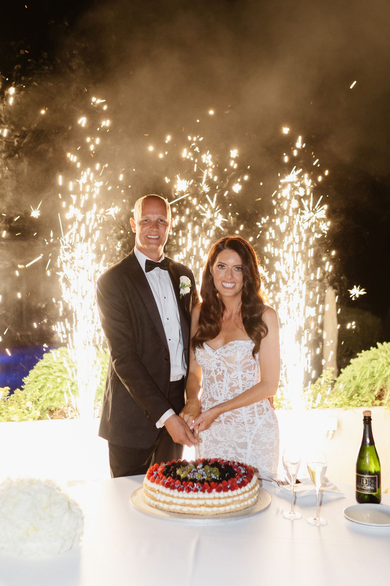Bride and groom cutting wedding cake with fireworks display behind them at elegant outdoor night reception.