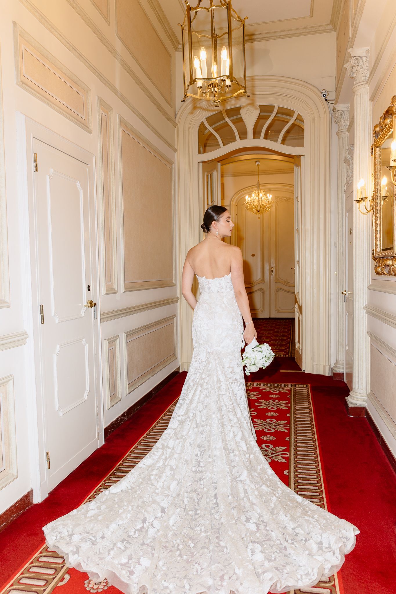 Bride in a stunning lace mermaid gown with long train walks through a luxurious hotel corridor.