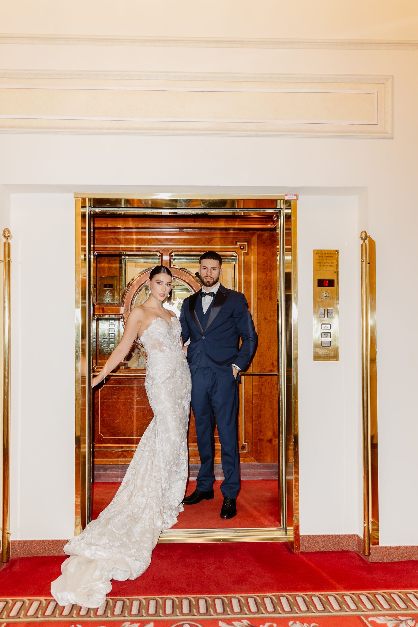 Bride and groom posing elegantly in a gold elevator, bride in lace gown, groom in navy suit with bow tie.