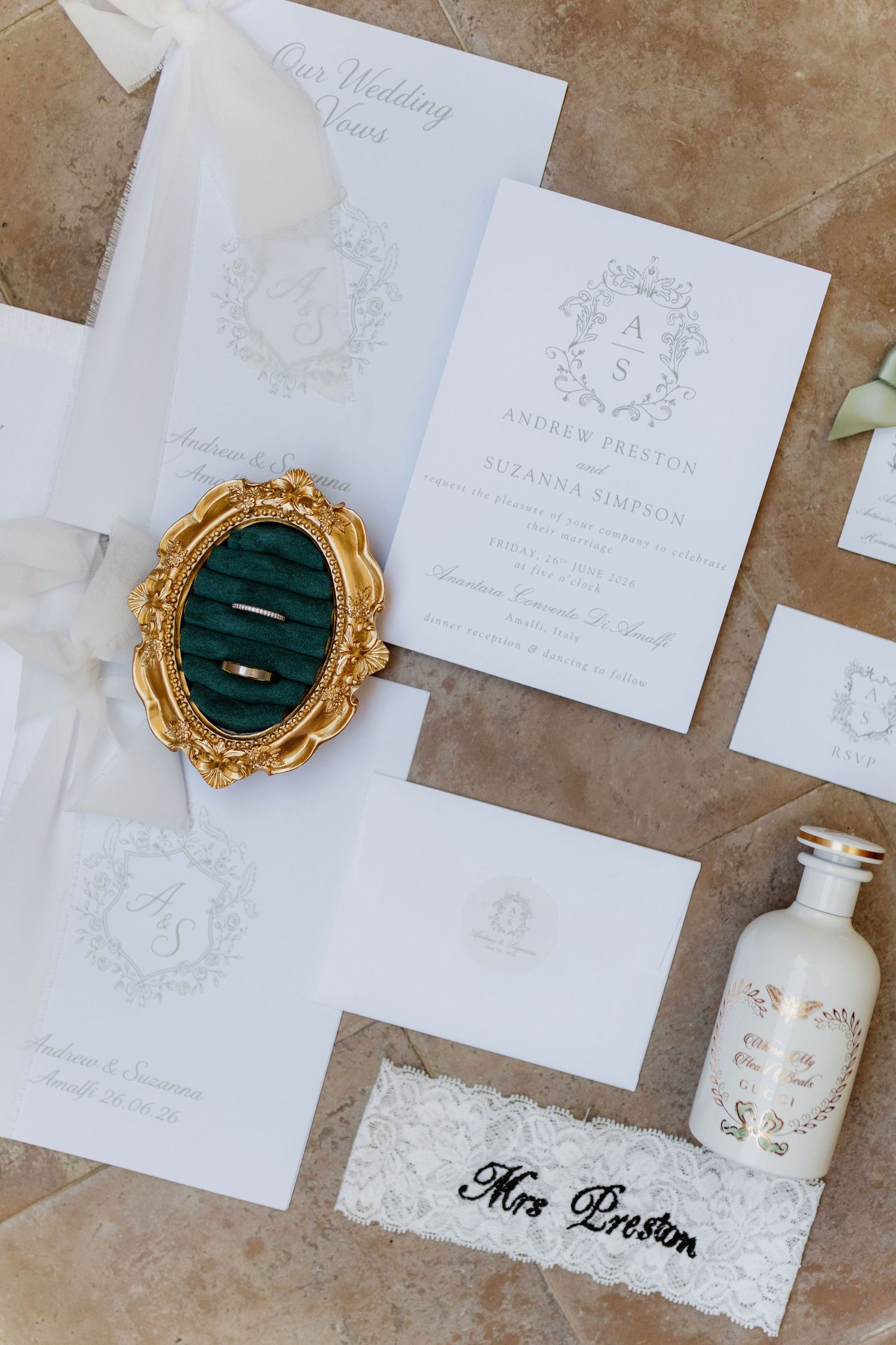 Elegant wedding stationery flat lay with monogram invites, gold ring holder, lace garter, and decorative bottle on tile.