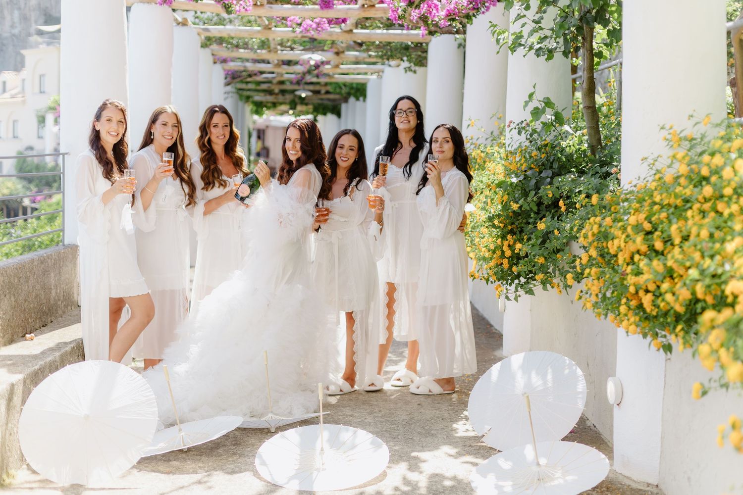 Bride and bridesmaids in white dresses celebrating on a sunlit terrace with columns and yellow flowers in Anantara Convento di Amalfi.