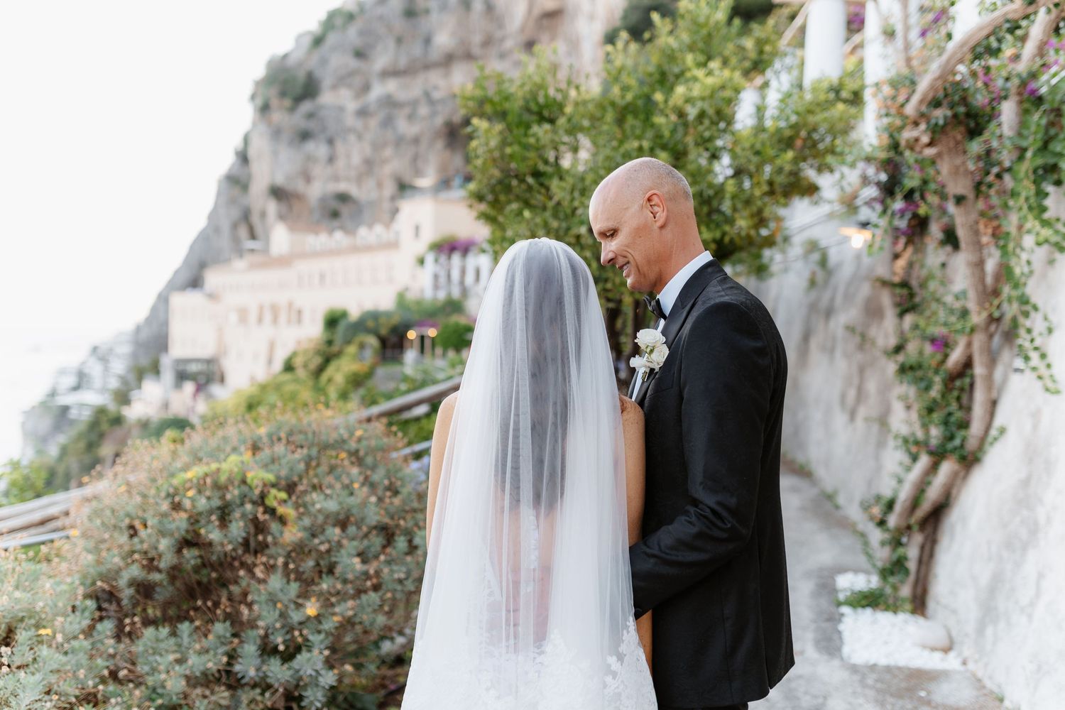 Bride and groom share a romantic first look moment on the Amalfi Coast, with cliffside villas and lush greenery behind them.