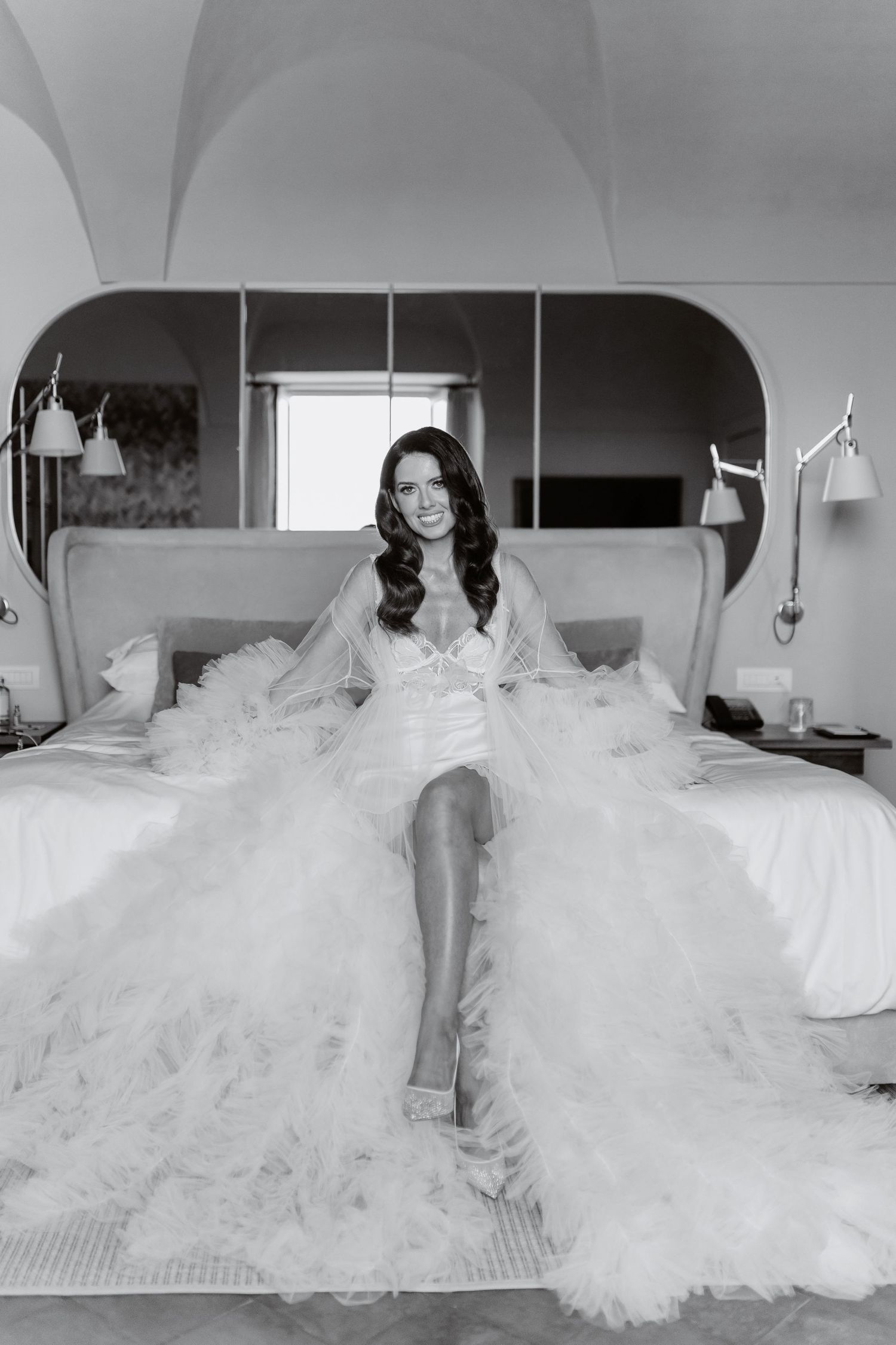 Black and white photo of a smiling bride in a ruffled wedding dress sitting on a bed in an elegant hotel room.