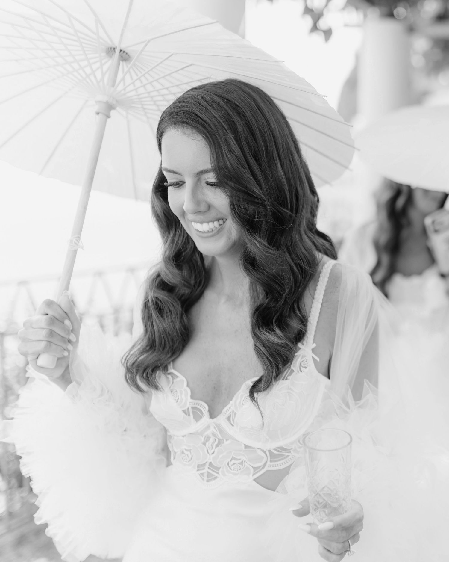 Smiling bride in lace gown holding a white parasol and champagne glass in a black and white photo.