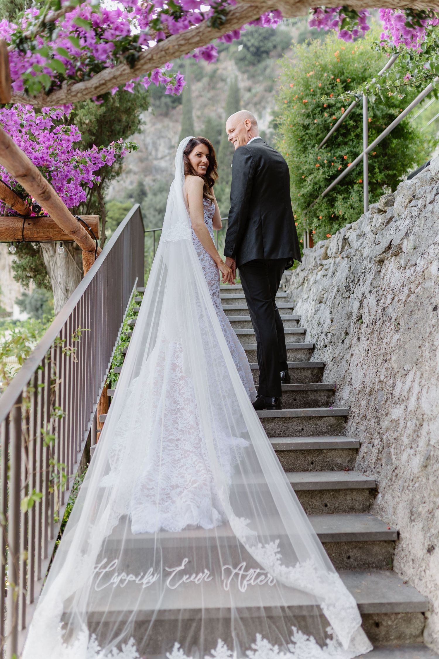 Bride in lace gown with cathedral veil holds hands with groom on stone steps surrounded by pink bougainvillea.