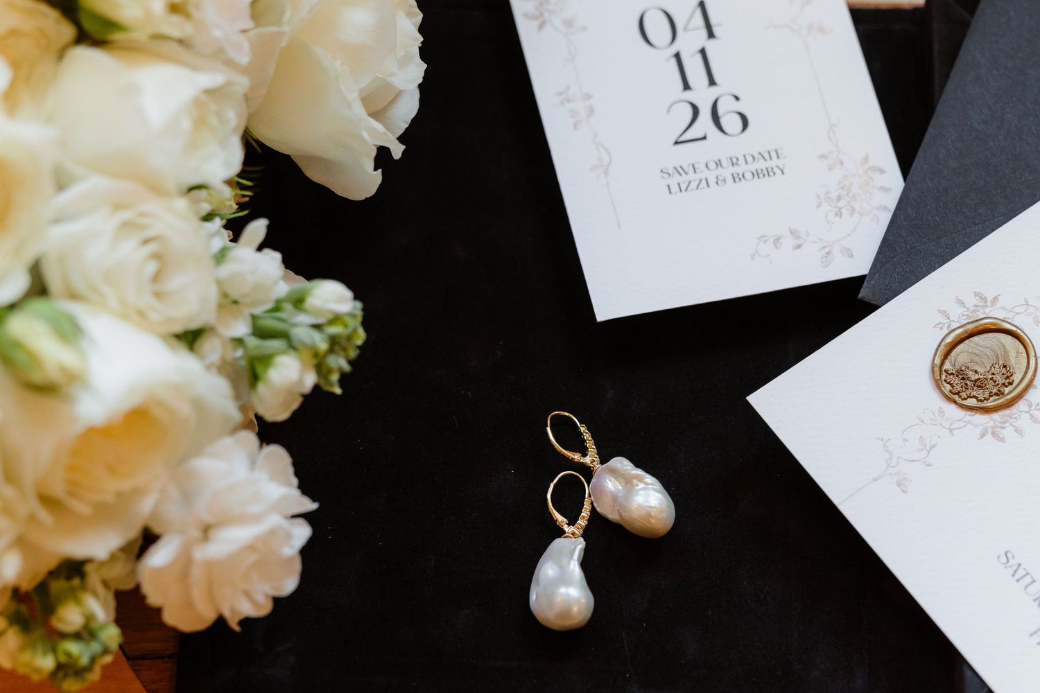 Wedding flat lay with pearl earrings, white roses, and elegant invitation dated 04.11.26.