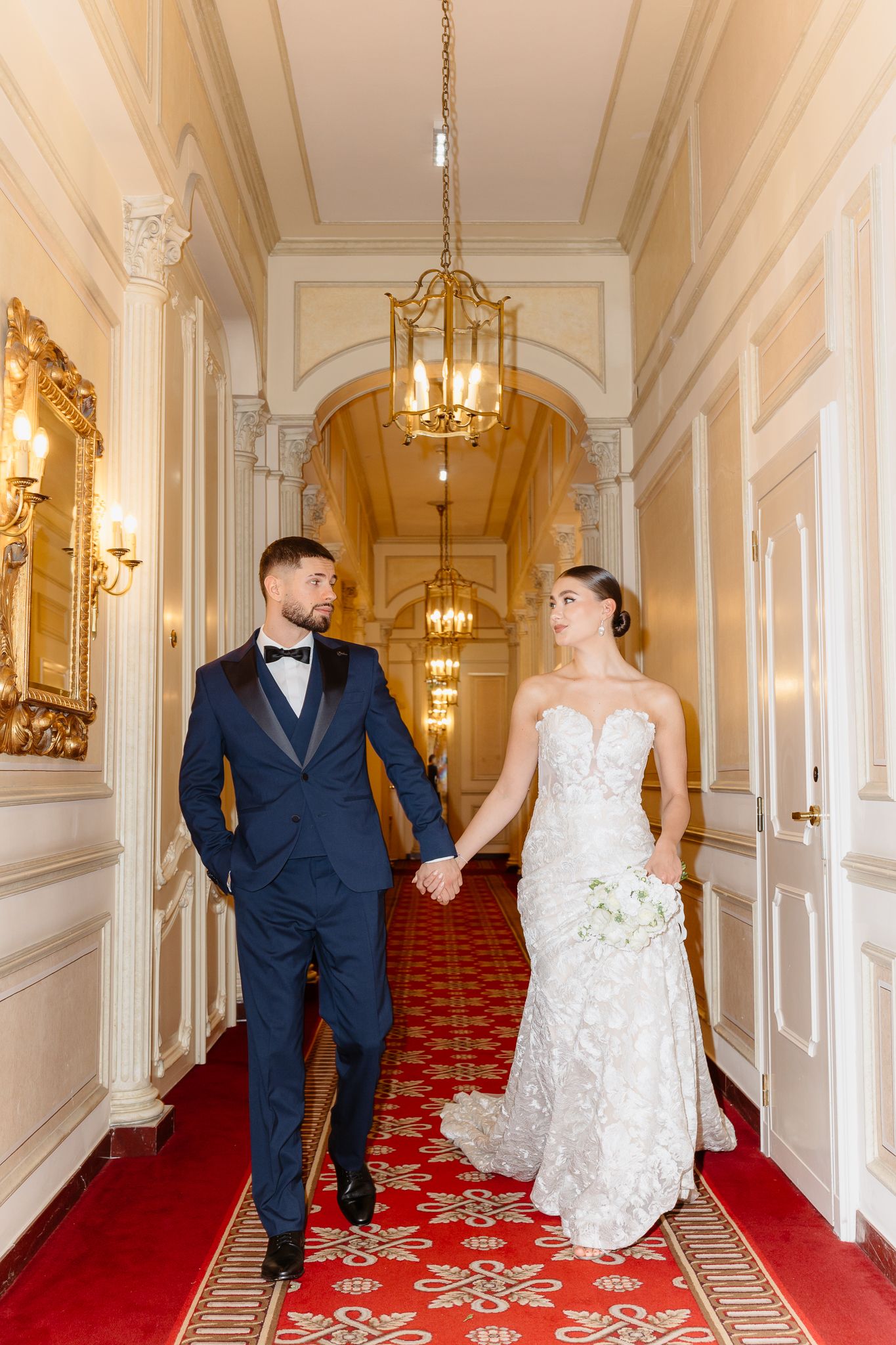 Bride and groom walk hand-in-hand through an elegant hallway with red carpet and gold chandeliers.