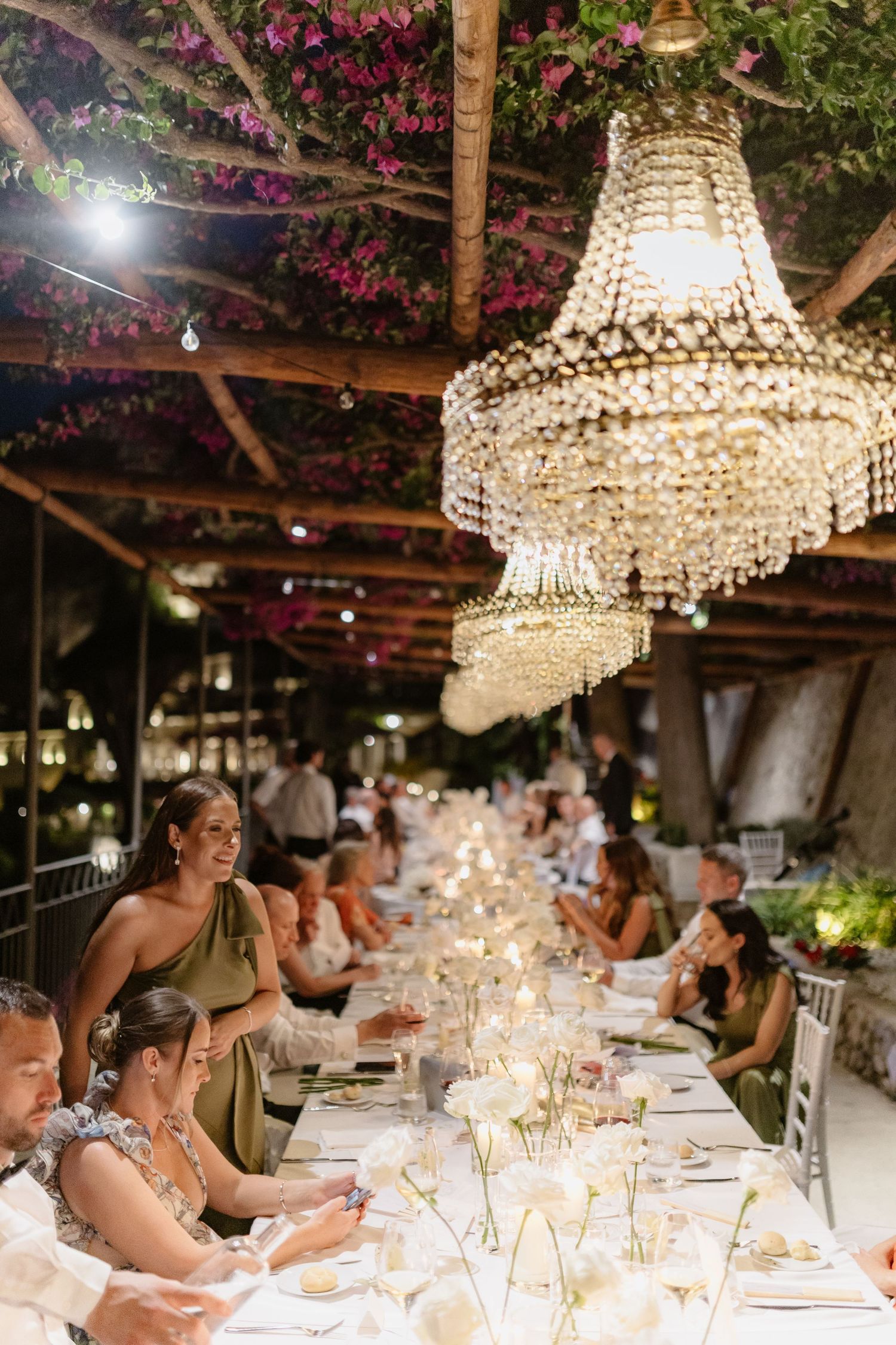 Elegant wedding reception with crystal chandelier, long dining table, floral ceiling decor, and guests in formal attire.