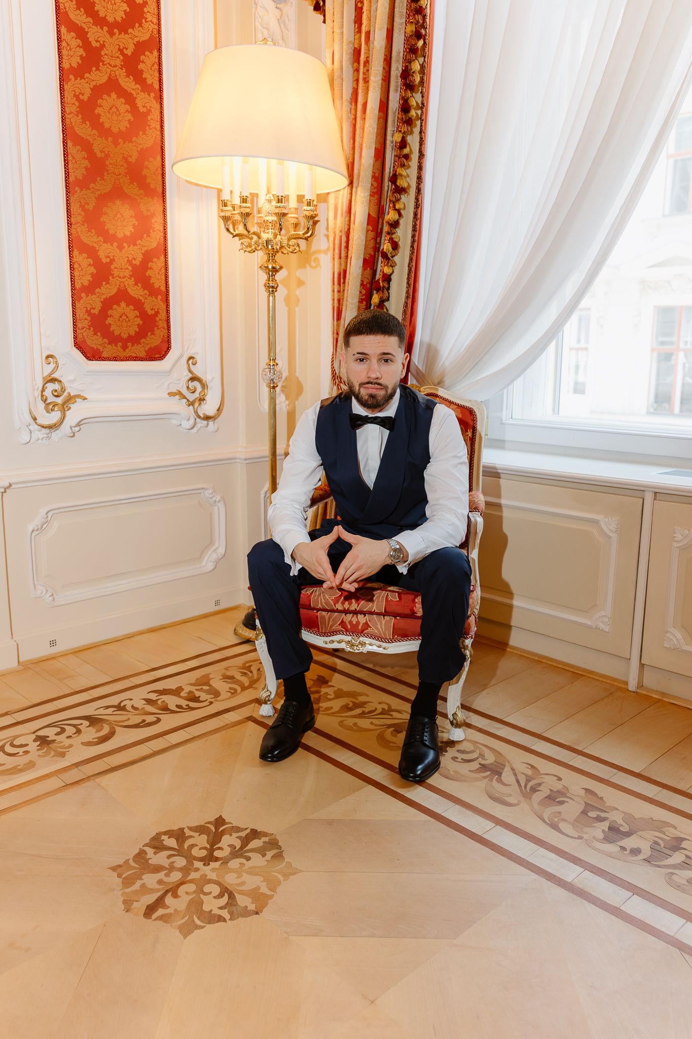 Dapper groom in navy vest and bow tie sitting in ornate red chair in luxurious hotel suite.