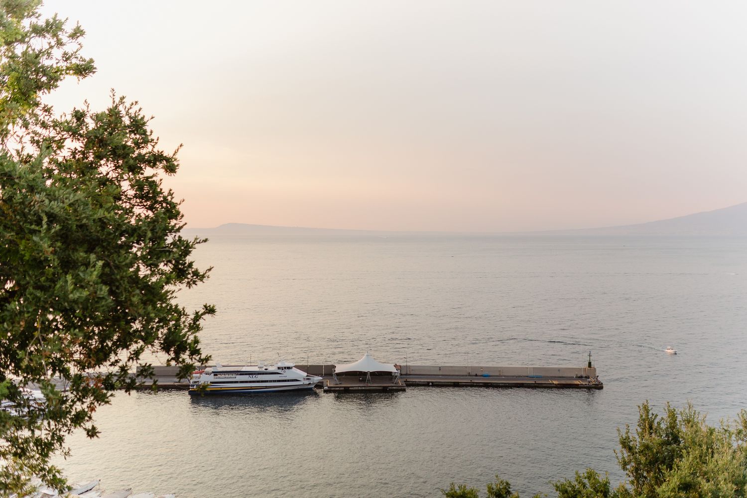 A serene dock with a boat moored at sunset over calm waters, framed by lush green trees and distant mountains.