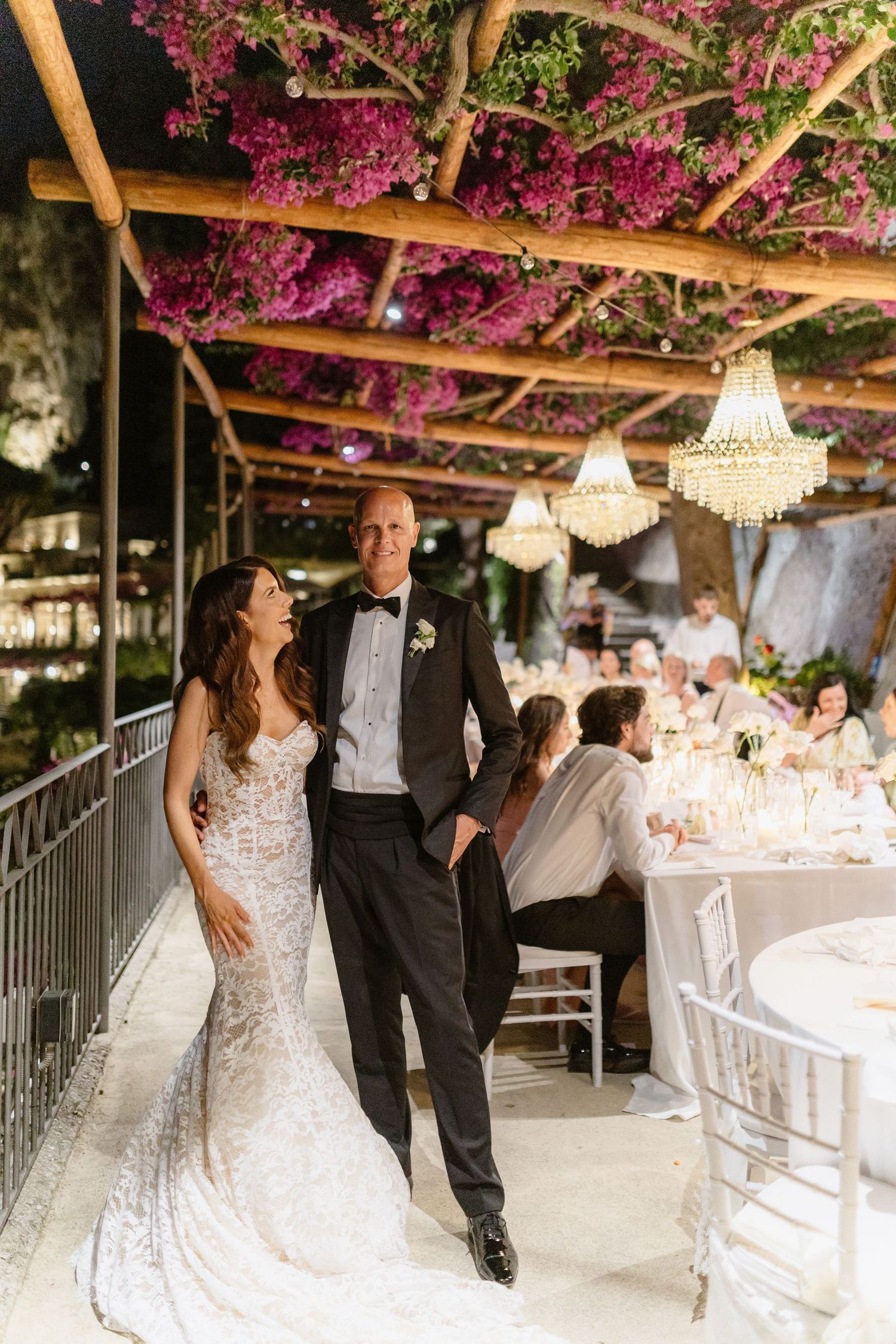 Bride and groom walk under bougainvillea pergola with chandeliers at elegant outdoor wedding reception.